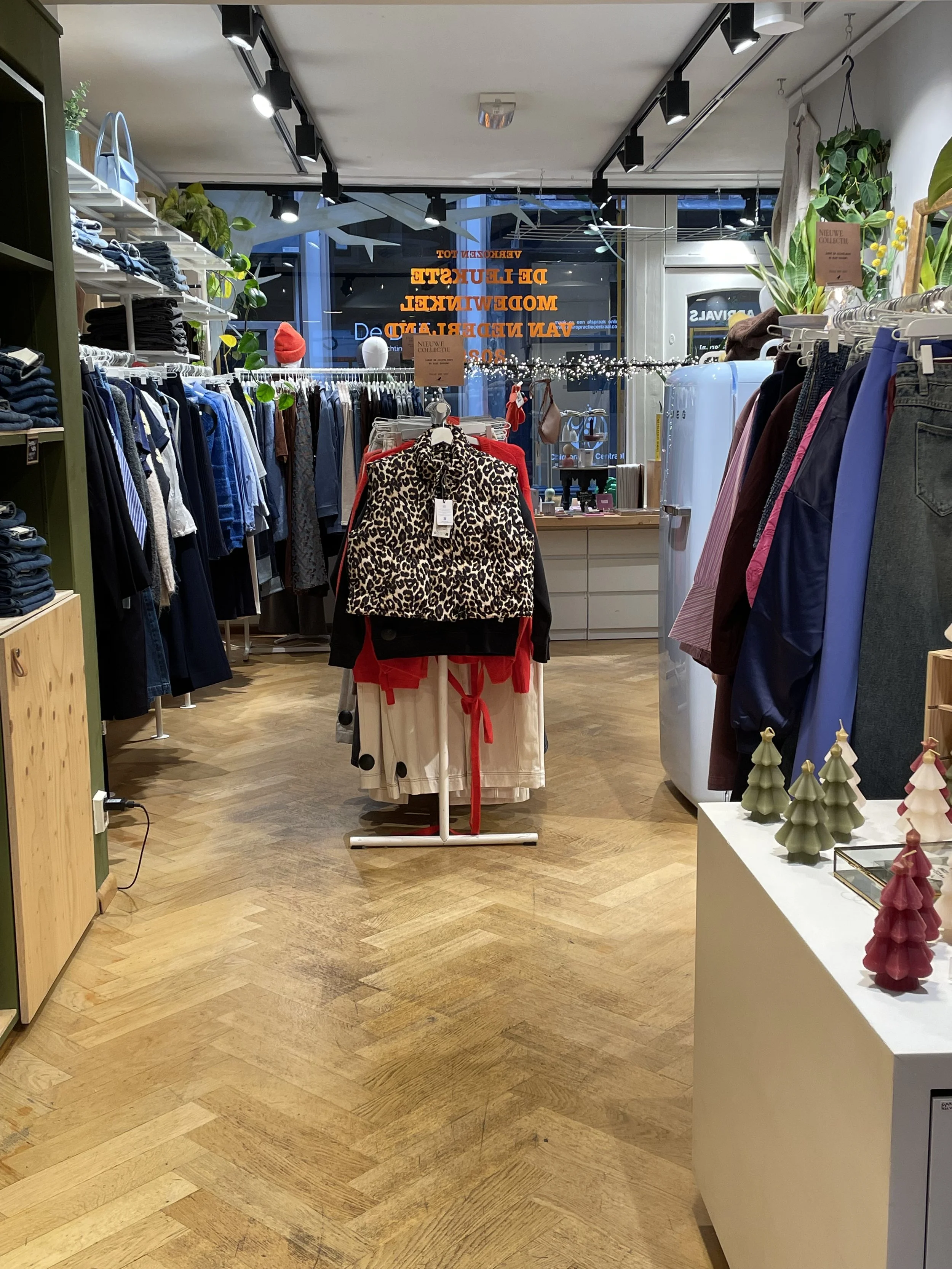Binnenkant van een kledingwinkel met rekken vol kleding, decoraties en een grote raampartij met tekst op glas.