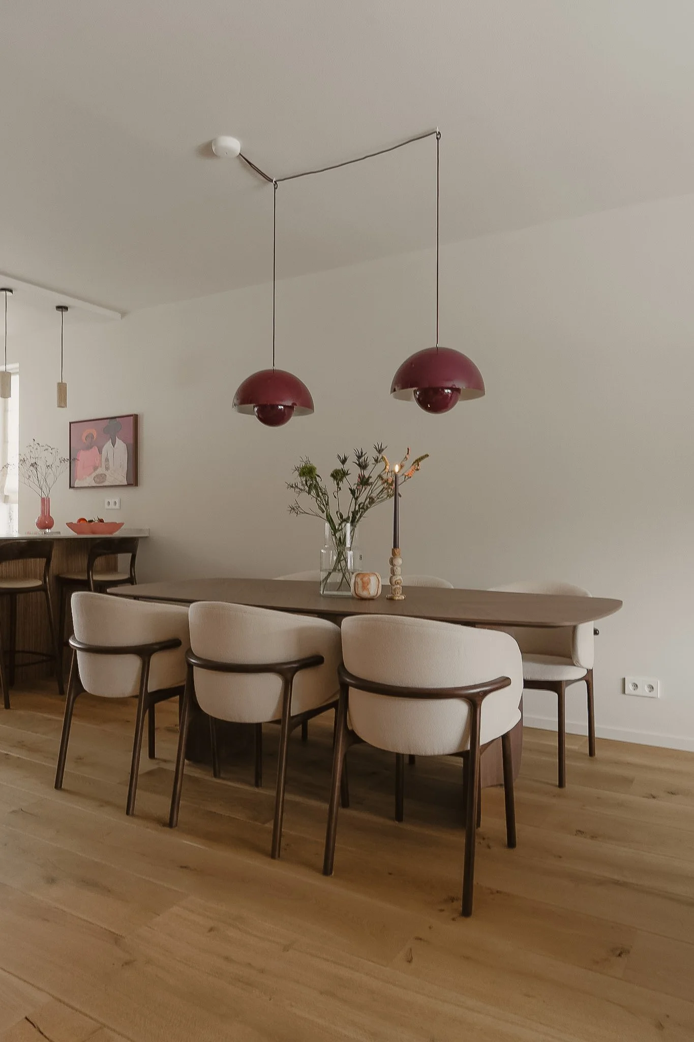 Een moderne eetkamer met een grote ovalen tafel en acht witte stoelen met houten poten. Boven de tafel hangen twee ronde, dieprode hanglampen. Op de tafel staat een vaas met bloemen en een paar decoratieve kaarsen. Op de muur achter de tafel hangt ee