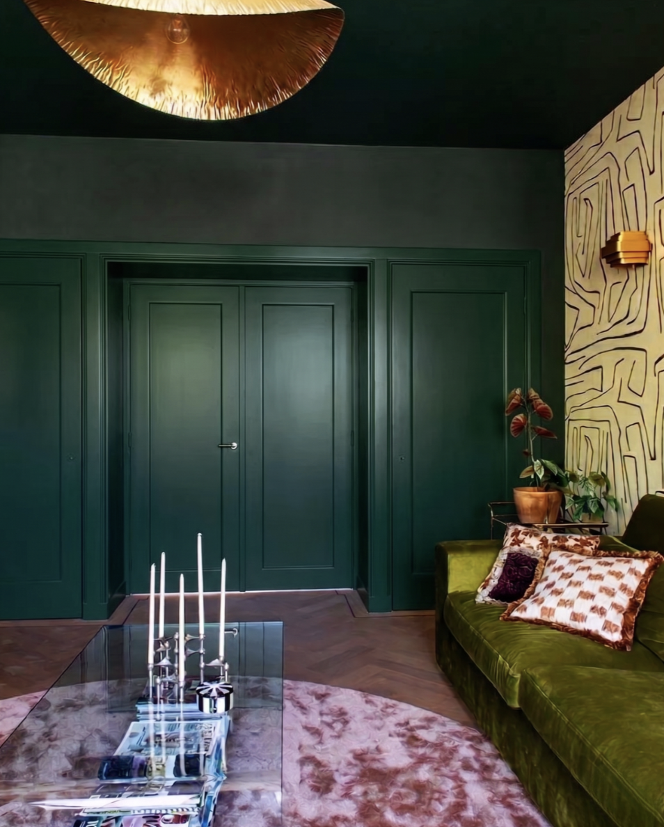 Een woonkamer met donkergroene deuren en muur, een groene bank met decoratieve kussens, een glazen salontafel met kandelaars, een rotan plantenpot met een plant, en een rozenhouten wand met een wandlamp. Een plafondlamp met een gouden afwerking hangt