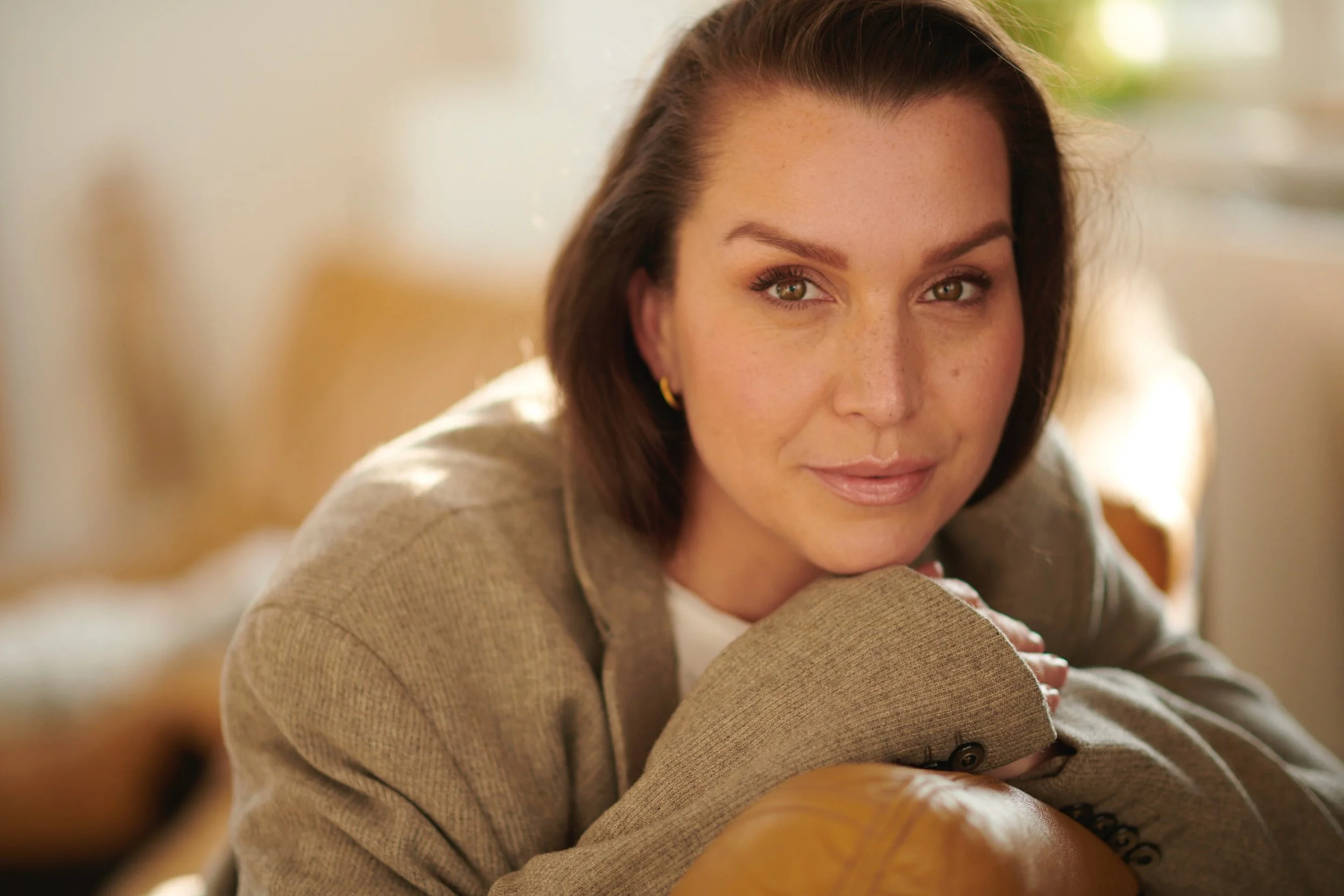 Person with short brown hair, wearing a tan suit jacket, resting head on hands, looking at the camera.