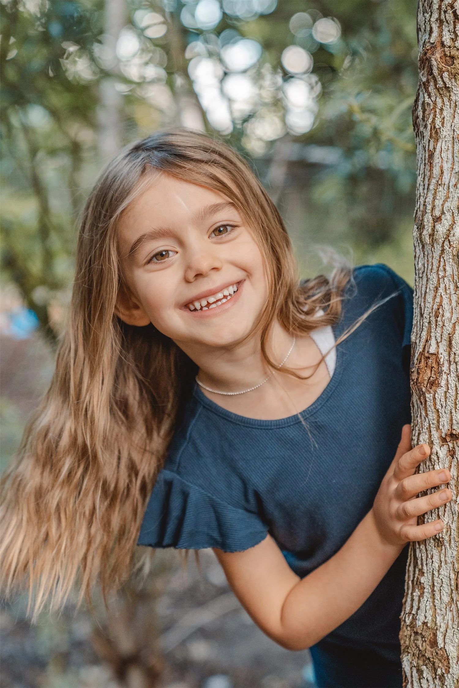 Ein Mädchen spielt mit der Kitafotografin „Kuckuck“. Das Mädchen im dunklen T-Shirt schaut lachend hinter einem Baum hervor und schaut in die Kamera.