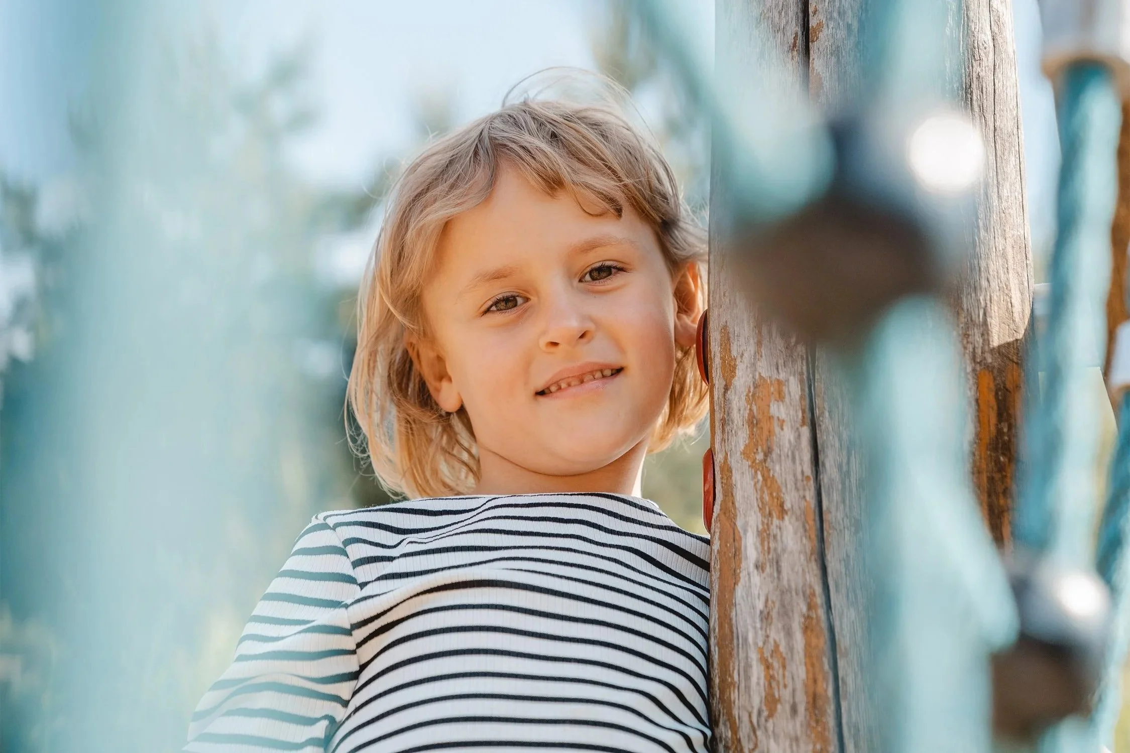 Kindergartenportrait. Blondes Mädchen steht auf einem Klettergerüst. Der Fotograf macht das Bild durch ein Kletternetz wodurch ein blauer Schimmer im Vordergrund entsteht.