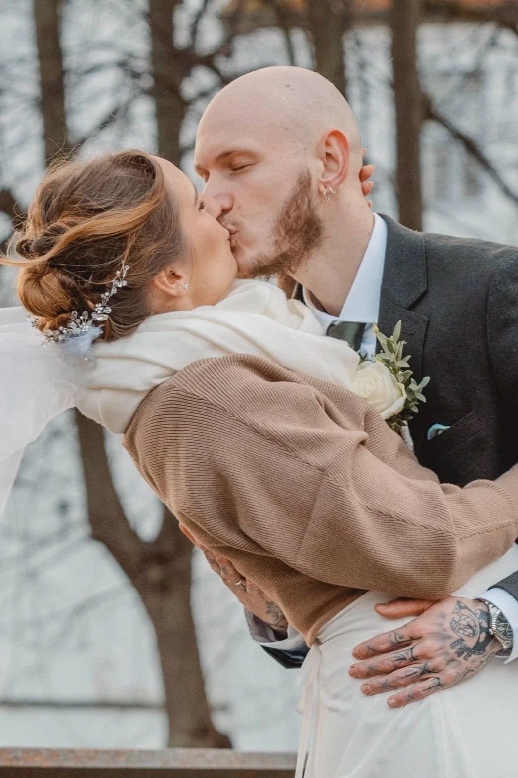 Hochzeitsportrait eines jungen Paares vor einem Schloss im Winter. Der Bräutigam hält seine Frau fest in den Armen und gibt ihr einen Kuss.  Die Braut trägt eine braune Stickjacke und einen weißen Schal über dem Kleid.