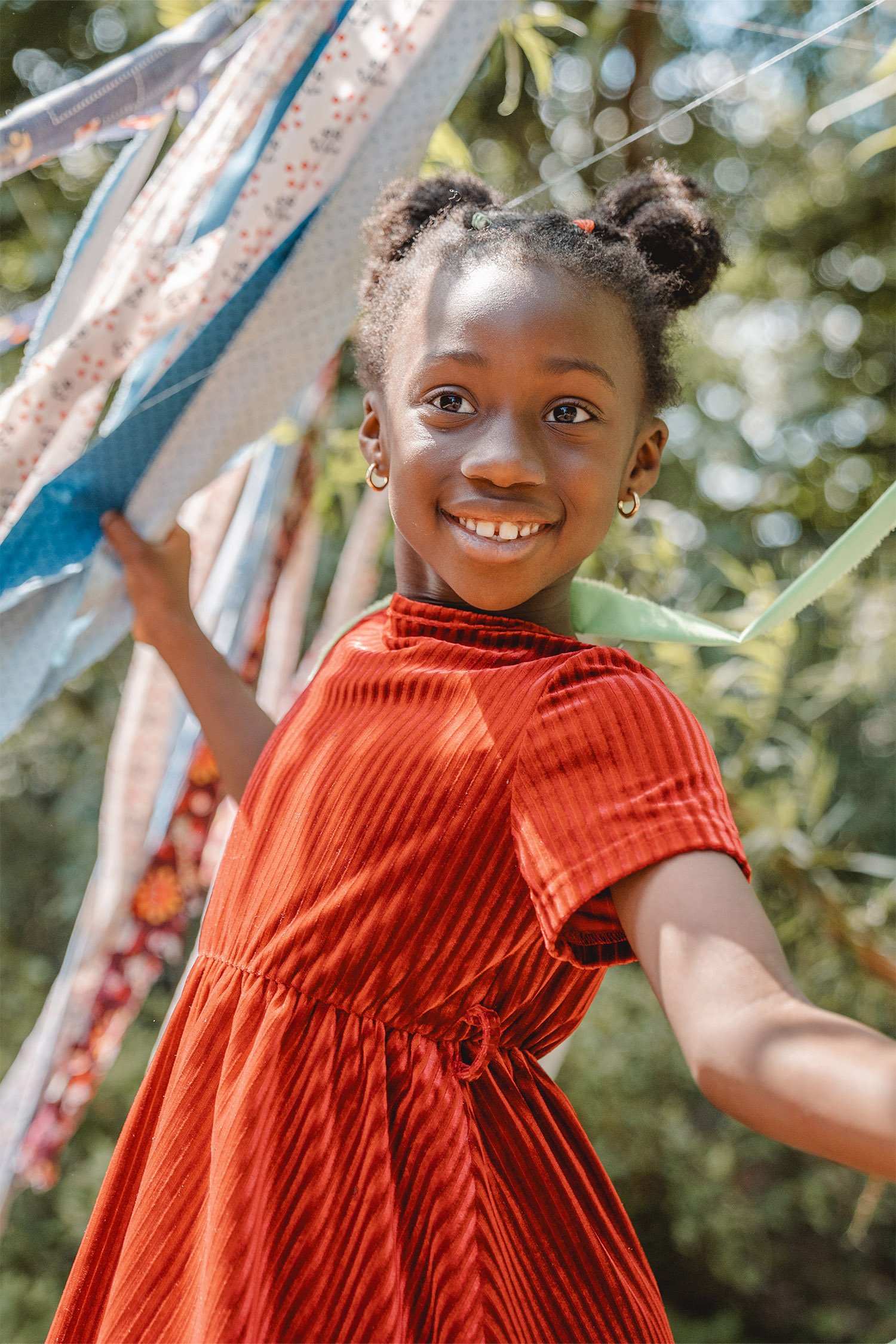 Kindergartenportrait von einem Mädchen in einem roten Kleid. Sie hält sich an bunten Tuchfetzen fest die von oben herab hängen. Sie lächelt und blickt an der Kamera vorbei in die Ferne.