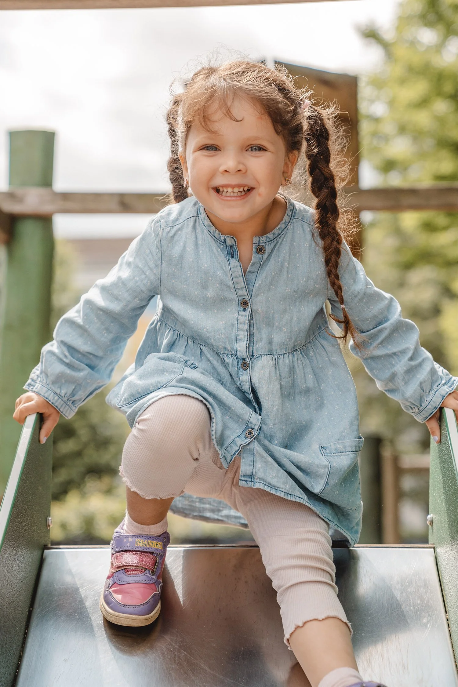 Outdoor Kitafoto von einem Mädchen mit zwei braunen geflochtenen Zöpfen. Sie setzt sich oben auf eine Rutsche und lächelt in die Kamera.
