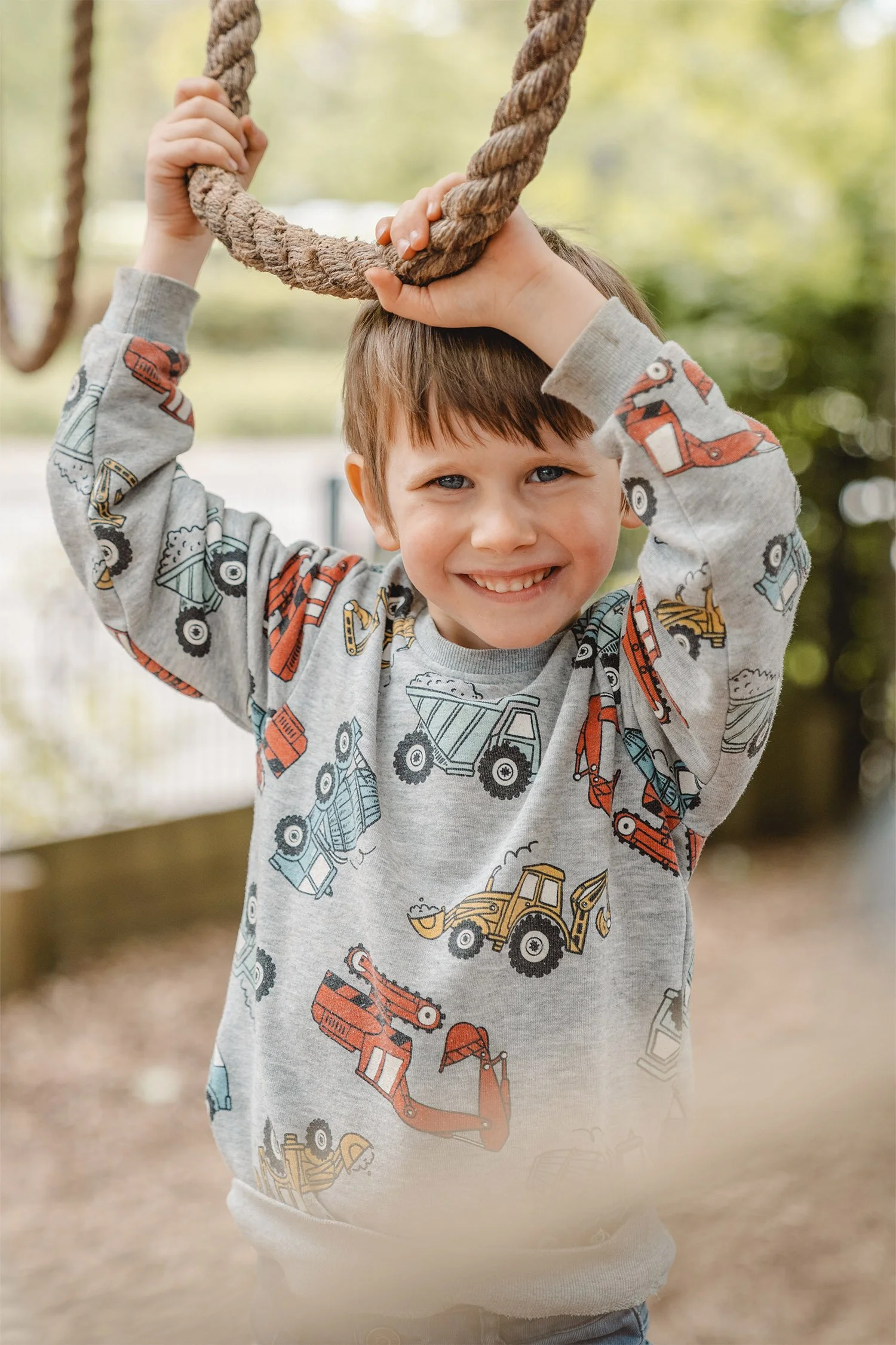Kitafoto von einem Jungen mit bunten Baggern auf dem Pullover. Er balanciert und hält sich über seinem Kopf an einem Tau fest. Dabei schaut und lächelt er in die Kamera.