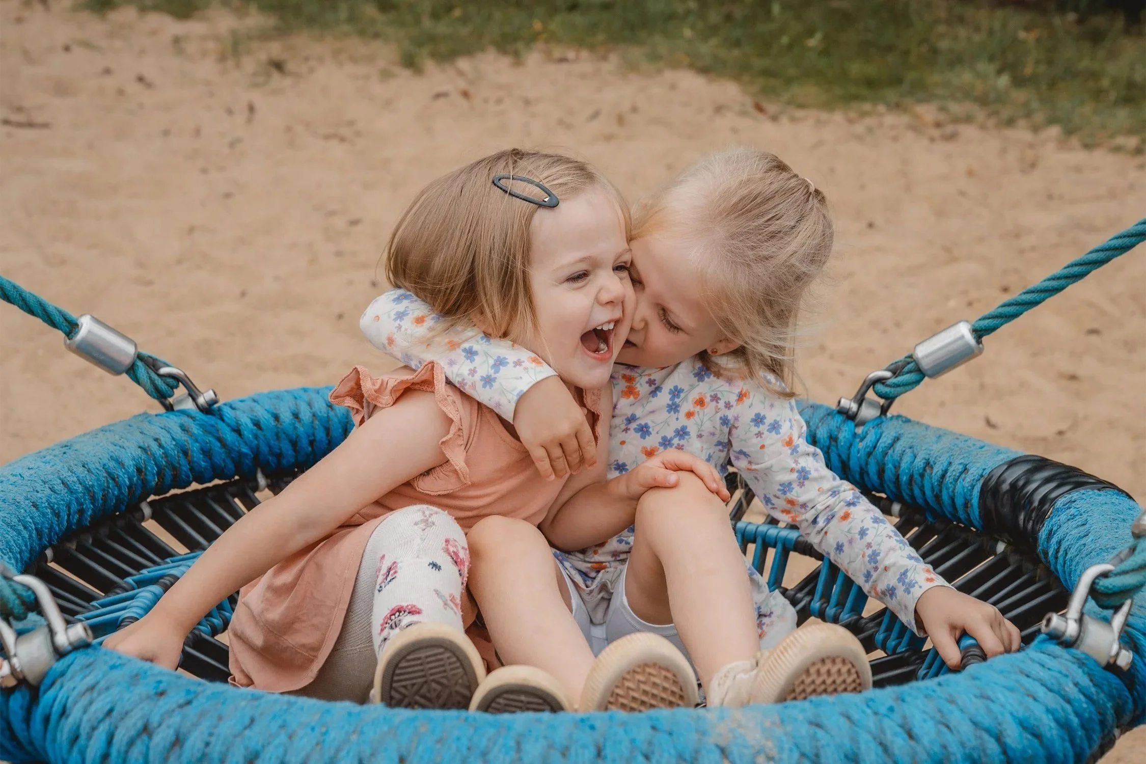 Geschwisterfoto im Kindergarten von zwei Schwestern die kuschelnd und lachend auf einer blauen Nestschaukel sitzen.
