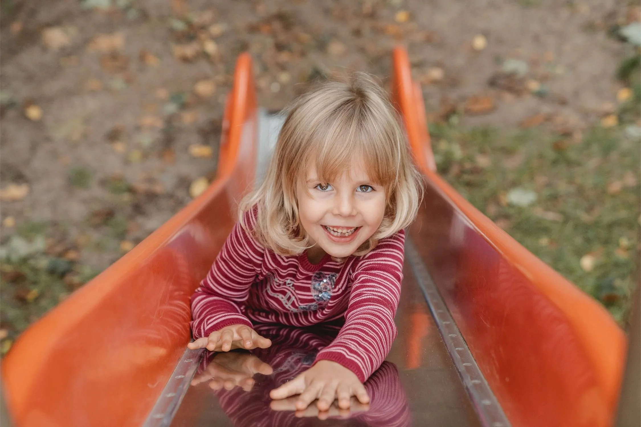 Ein Mädchen rutscht beim Kitashooting die Rutsche auf dem Bauch hinunter und schaut dabei lachend nach oben zur Fotografin, die oben an der Rutsche steht. 