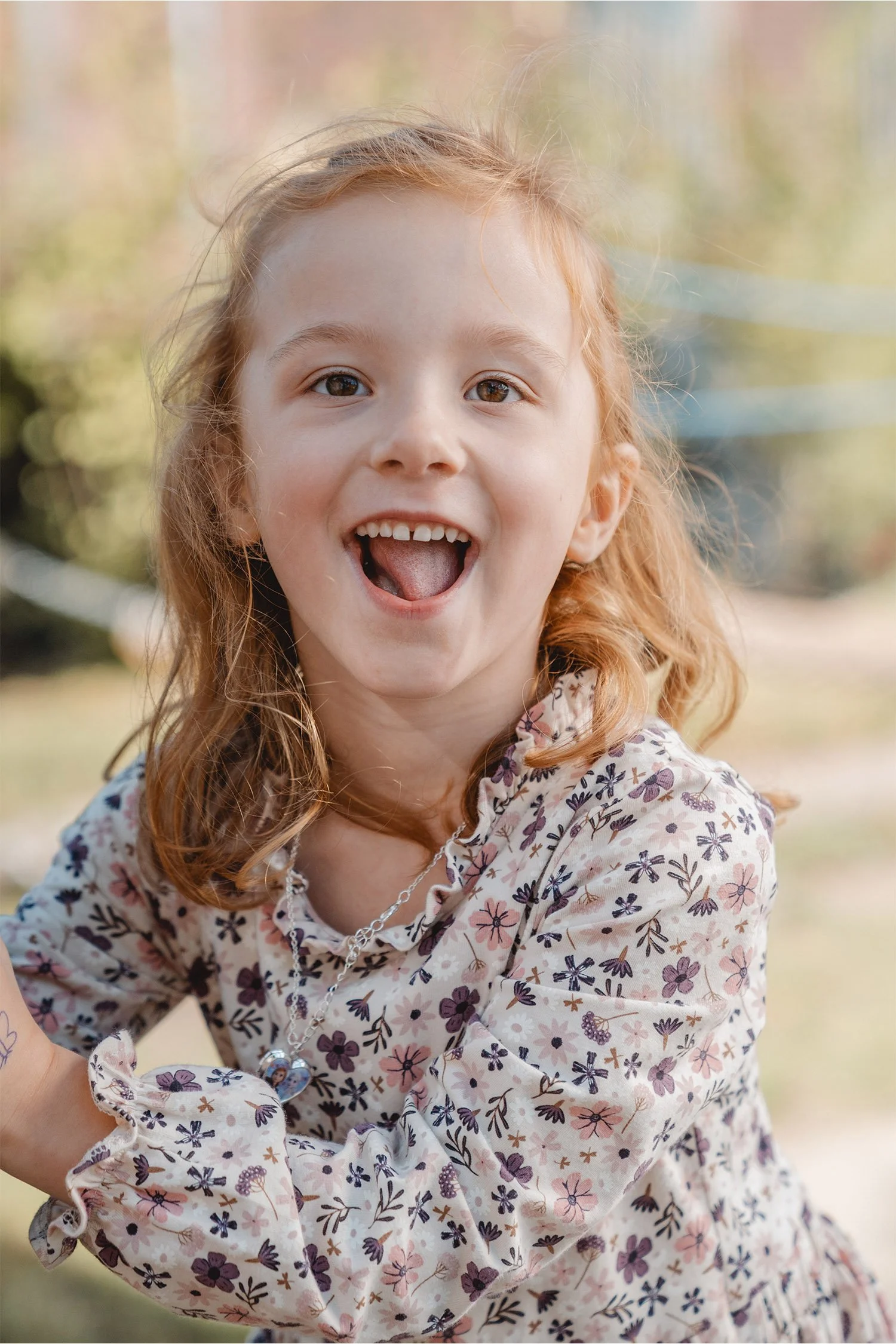 Ein Mädchen mit roten Haaren und einem weißen Blümchenkleid schaut am Kita-Fototag glücklich und lachend in die Kamera des Fotografen.