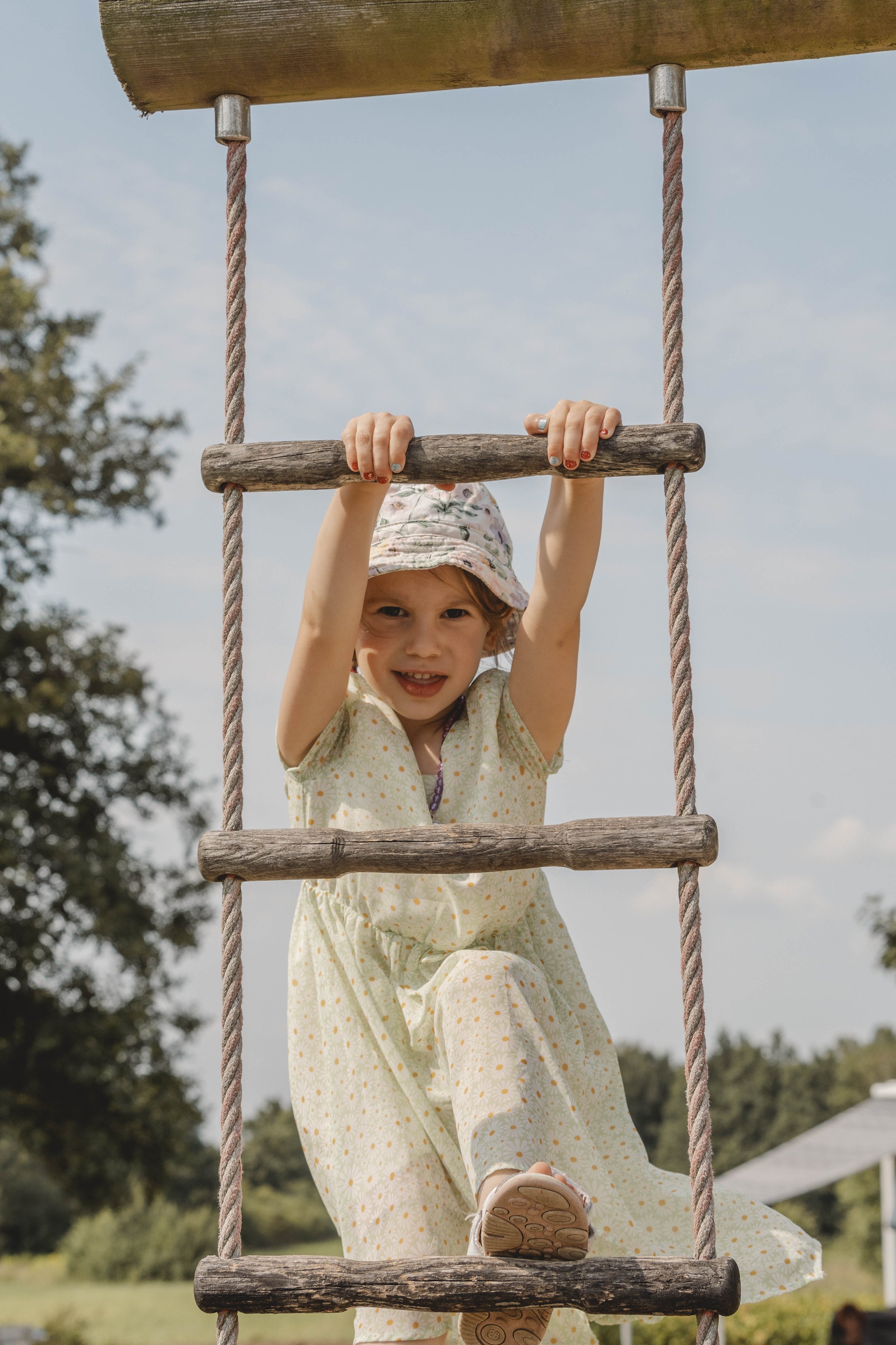 Frontal zentriertes Kitafoto von einem Mädchen mit Hut und einem hellgrünen Kleid. Sie schaut in die Kamera und klettert eine Strickleiter hoch.