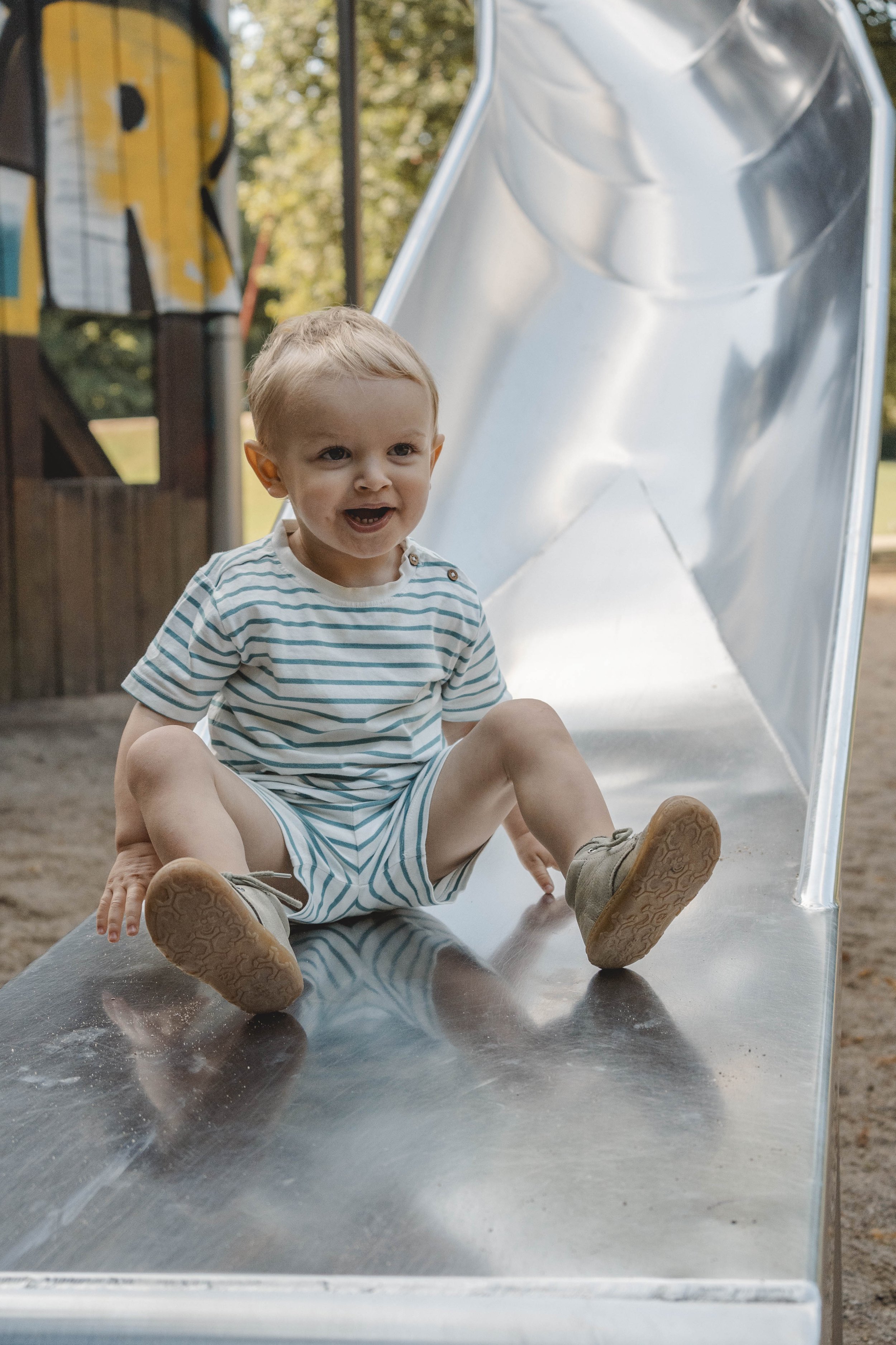 Kindergartenfoto von einem Jungen mit blau-weiß gestreiftem T-Shirt und kurzer Hose. Er sitzt am Ende einer großen Rutsche aus Aluminium. 