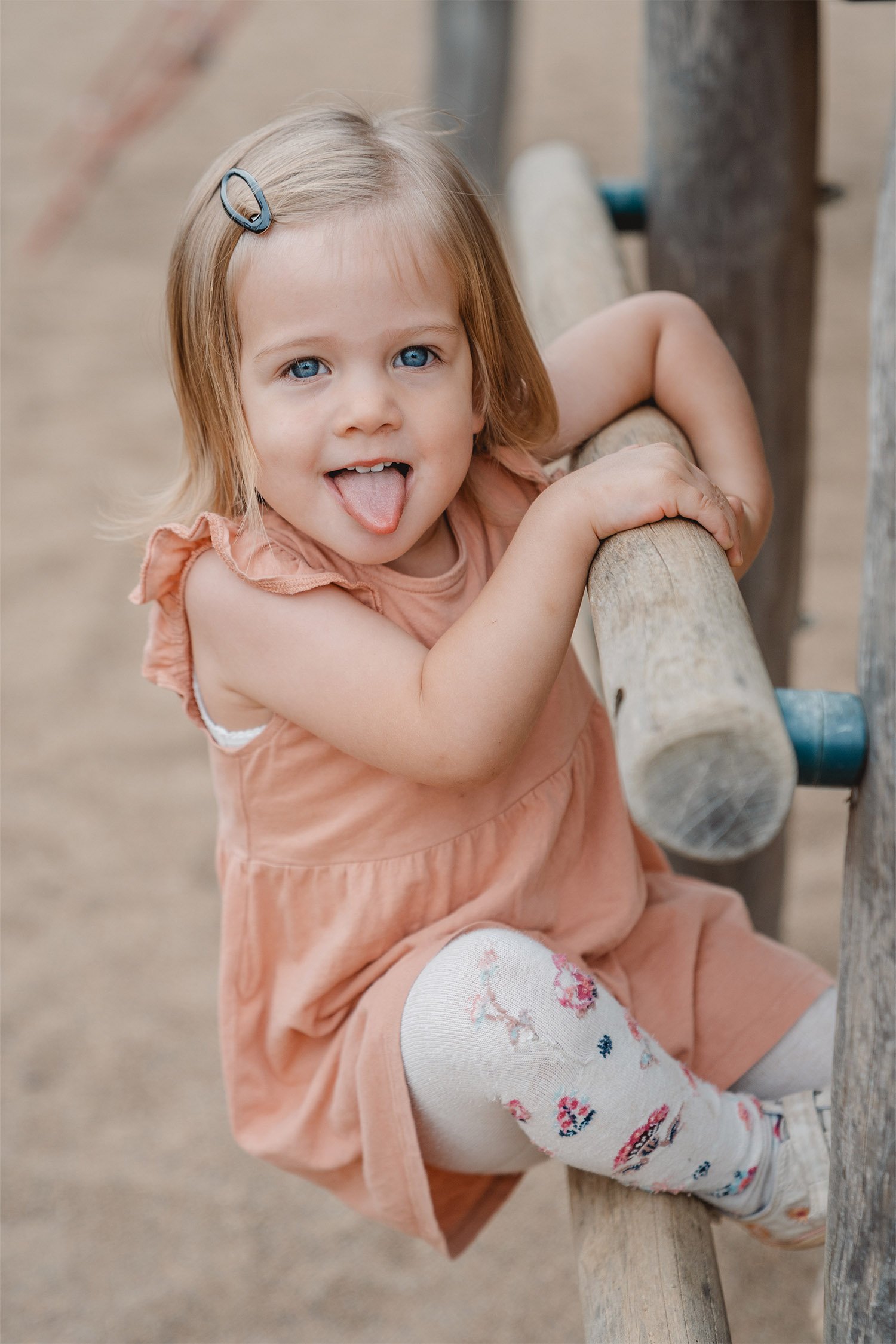 Kindergartenfoto von einem Mädchen in einem rosafarbenen Kleid. Sie klettert ein Klettergerüst hoch, schaut in die Kamera und streckt ihre Zunge heraus. 