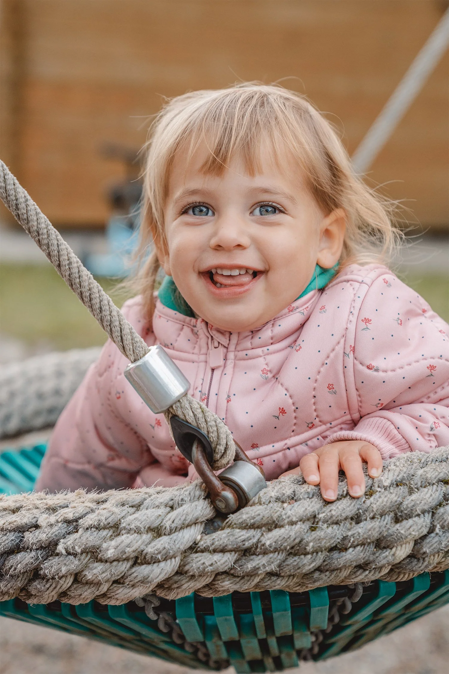Krippenkind in einer rosafarbenen Jacke sitzt in einer Nestschaukel und schaut lachend in die Kamera.