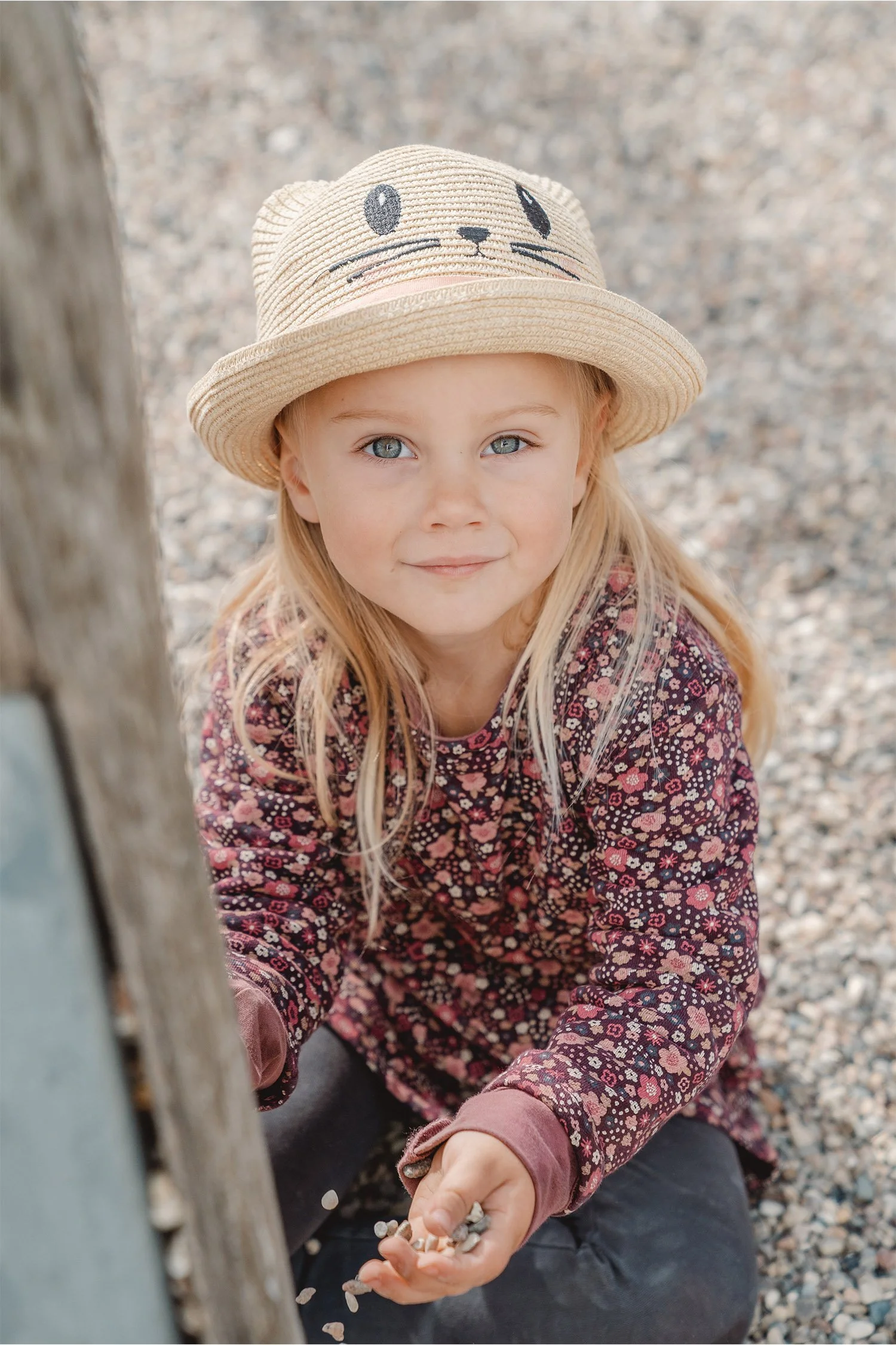Kindergartenportrait von einem blonden Mädchen, das im Kiessand sitzt. Sie trägt einen Strohhut und spielt mit Steinchen. 