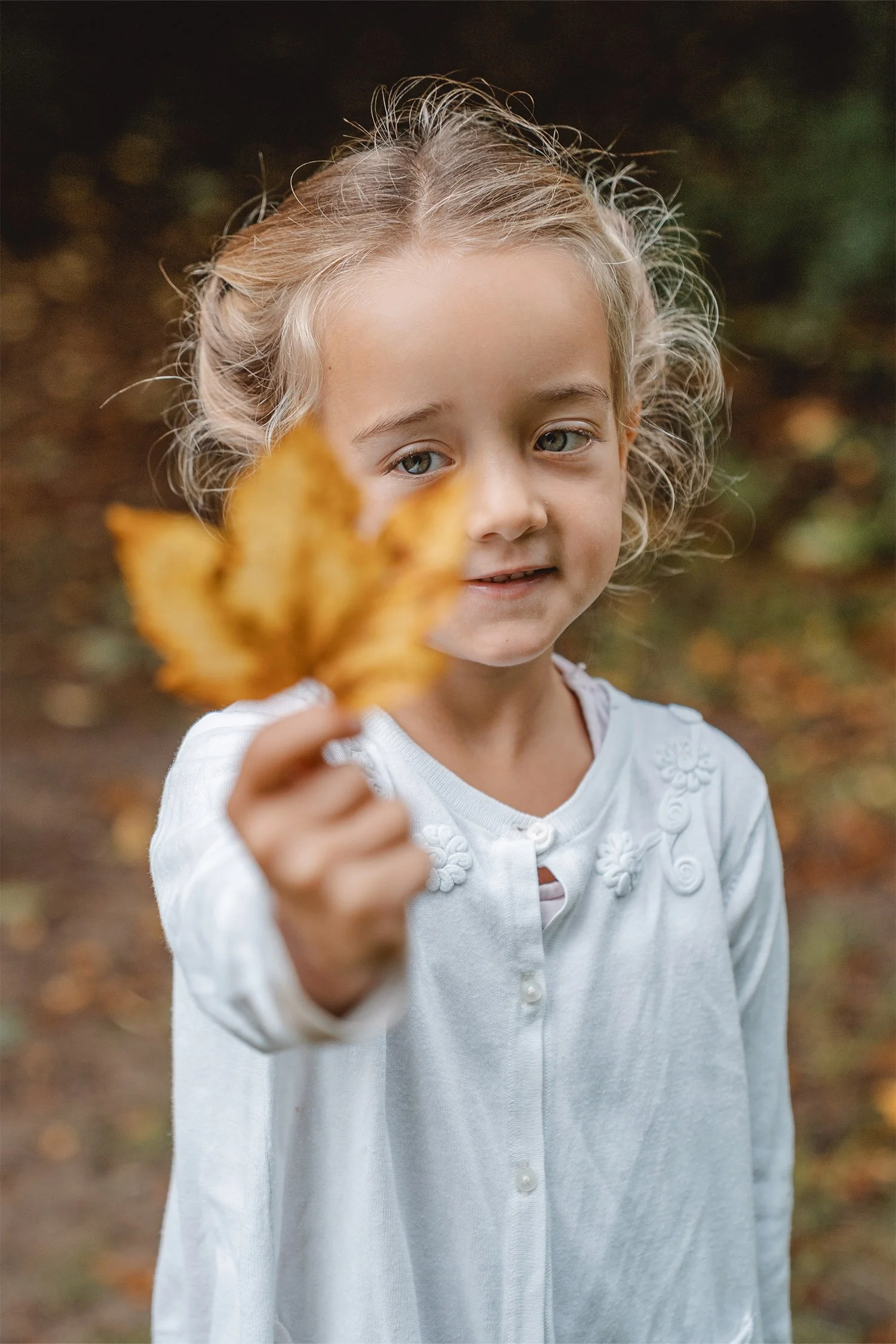 Ein blondes Mädchen hält beim Kita-Herbstshooting ein gelbes Ahornblatt in die Kamera. Sie schaut sich das Batt an. Ihre Augen sind im Fokus. Das Blatt und ihre Hand sind unscharf. 