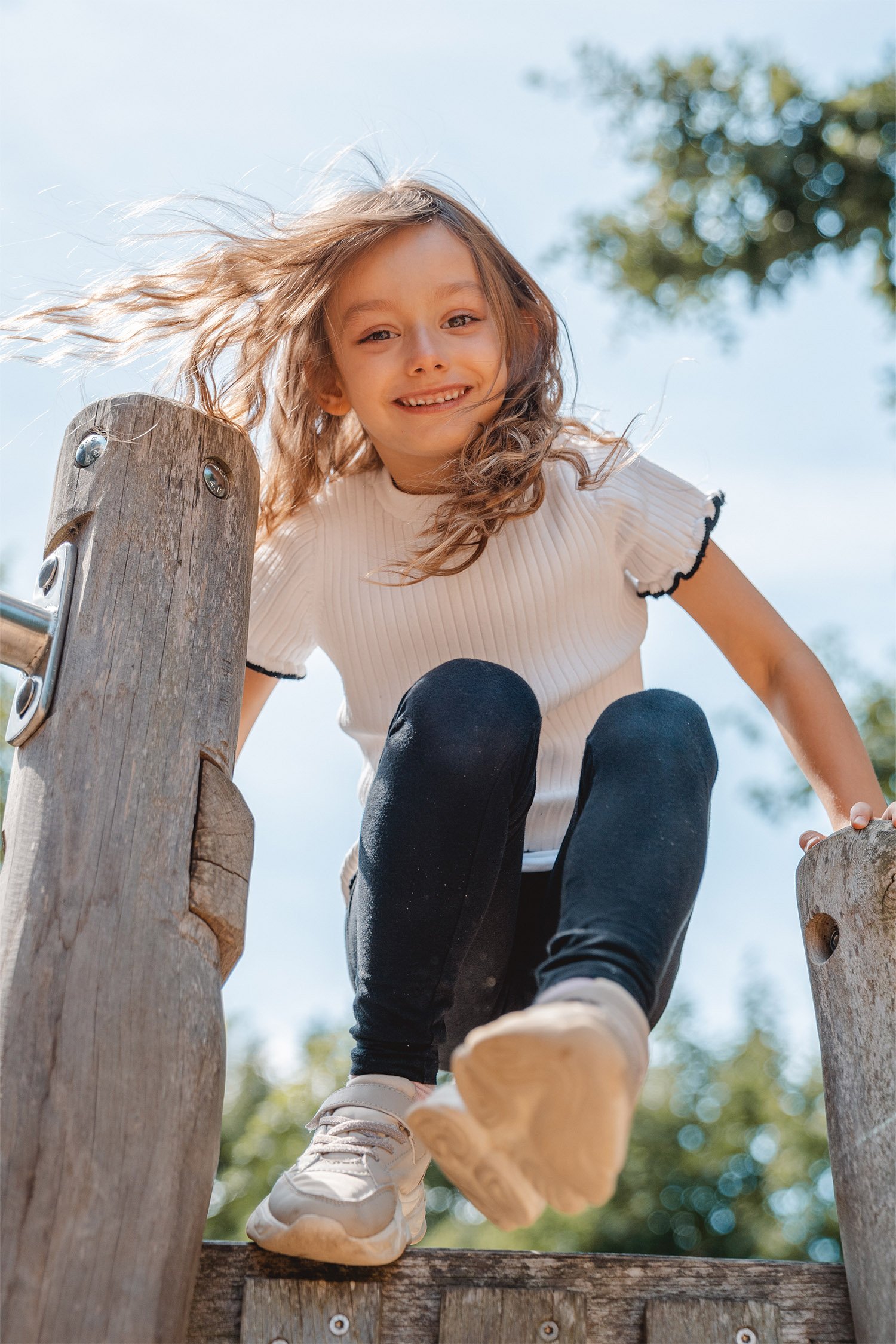 Ein Mädchen hopst mit Schwung beim Outdoor-Kitashooting über ein Geländer vom Klettergerüst und schaut dabei lächelnd in die Kamera.