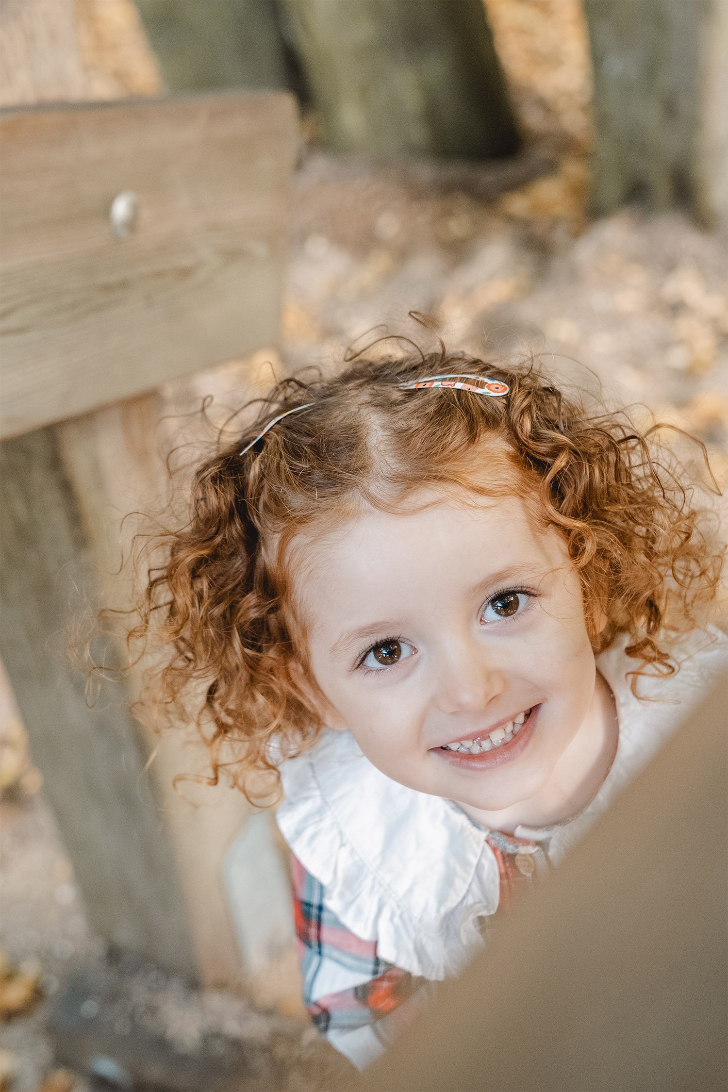 Ein von oben fotografiertes Mädchen mit rot gelockten Haaren klettert eine Treppe zum Klettergerüst hoch. Sie schaut lächelnd in die Kamera. Im Vordergrund sieht man verschwommen einen Teil vom Geländer.