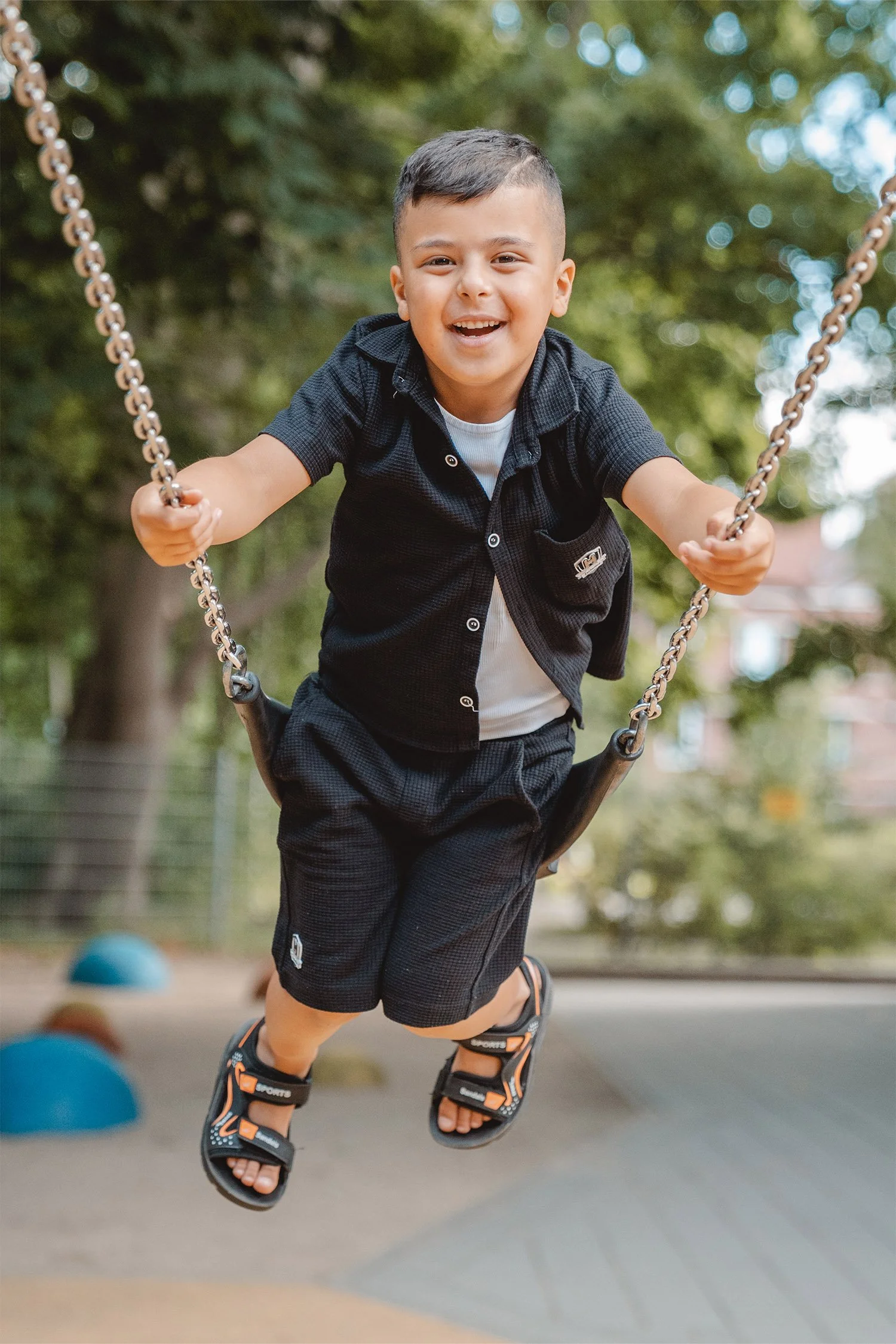 Ein Junge schaukelt beim Kitashooting sehr hoch und lacht dabei in die Kamera. Er trägt Hemd, Hose und Sandalen in schwarz.