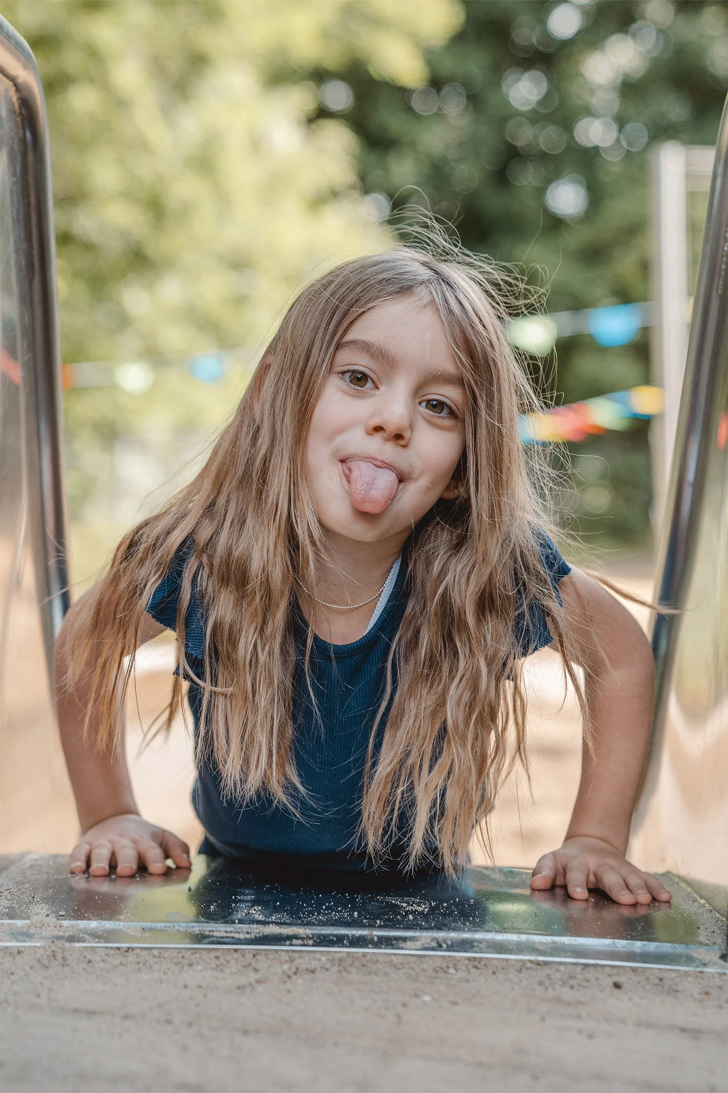 Ein Mädchen mit brauen langen Haaren und blauem T-Shirt liegt mit ihrem Bauch oben auf der Rutsche und streckt fröhlich ihre Zunge in Richtung des Fotografen beim Kitashooting.