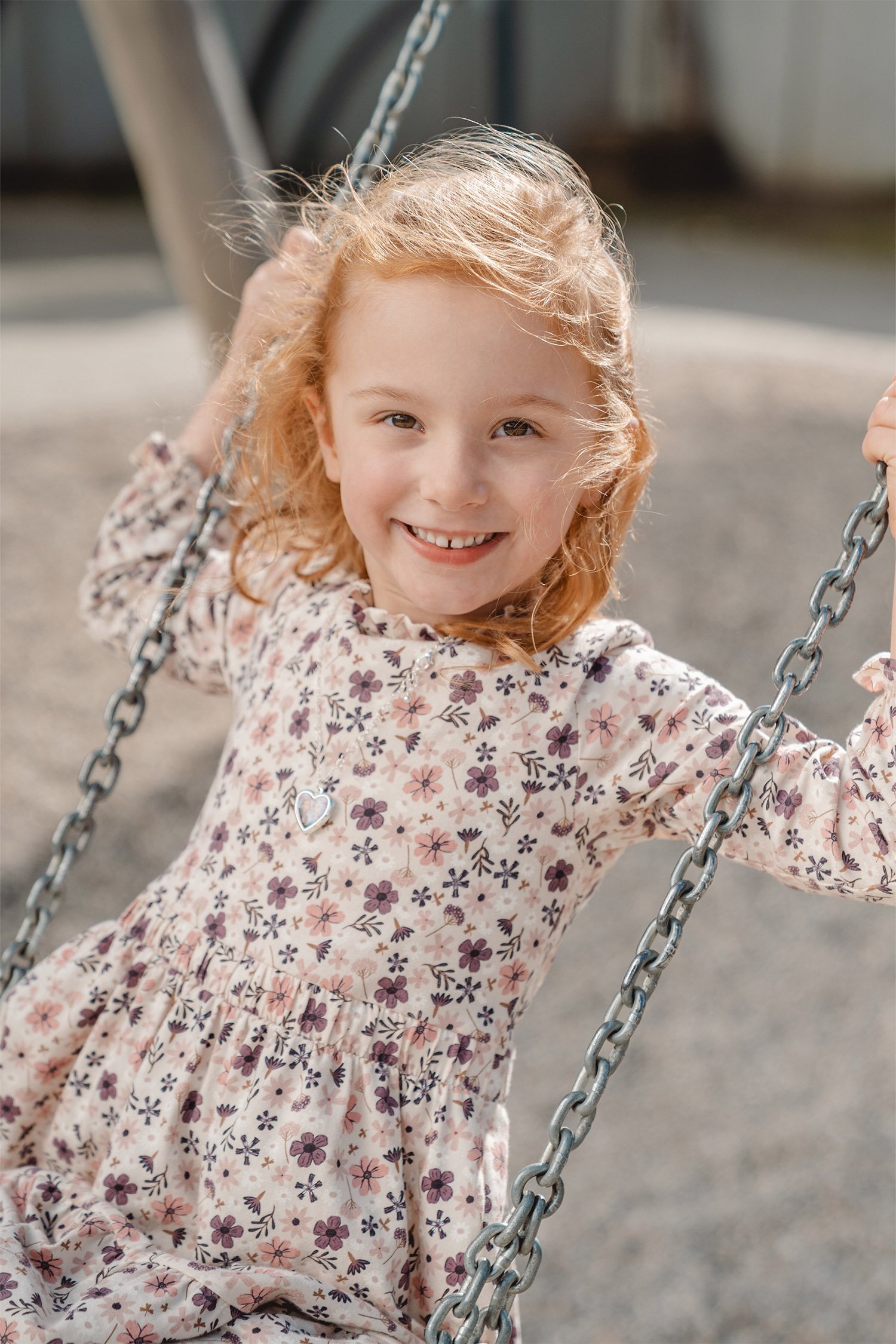 Ein Kitafoto von einem Mädchen in einem Blümchenkleid auf einer Schaukel. Das Bild ist von der Seite gemacht. Das Mädchen hat rote Haare und lächelt in die Kamera. 