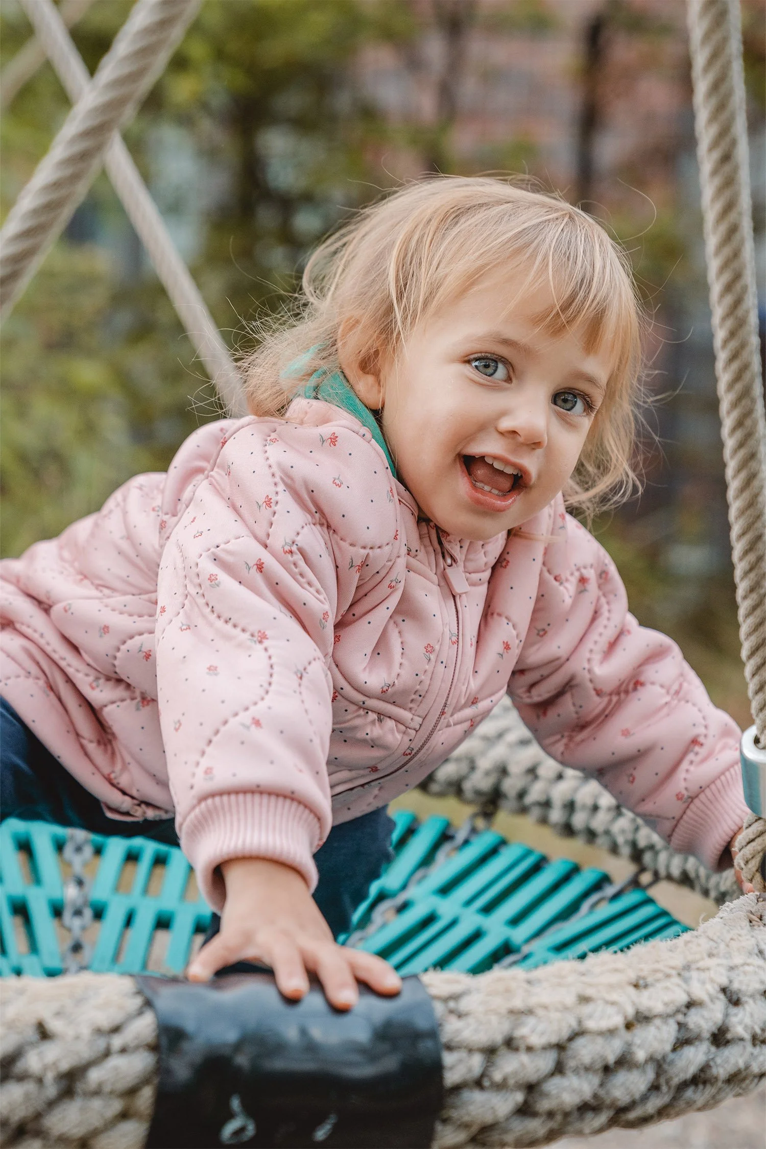 Ein Krippenkind versucht auf einer Nestschaukel aufzustehen und hält sich dabei gut fest. Sie lacht und schaut dabei in die Kamera. 