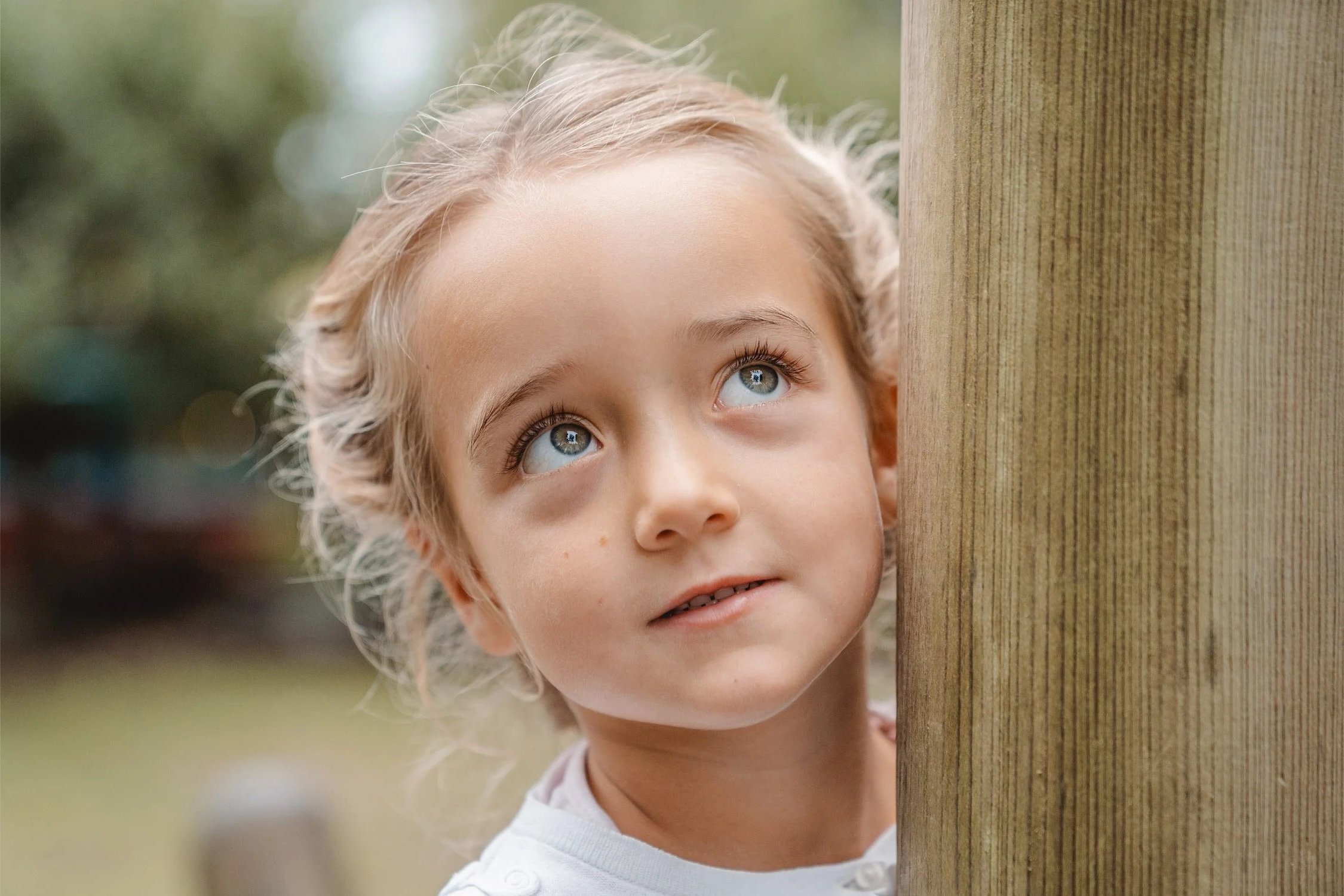 Portrait von einem blonden Mädchen. Sie steht an einem Holzpfahl und schaut nachdenklich nach rechts oben.