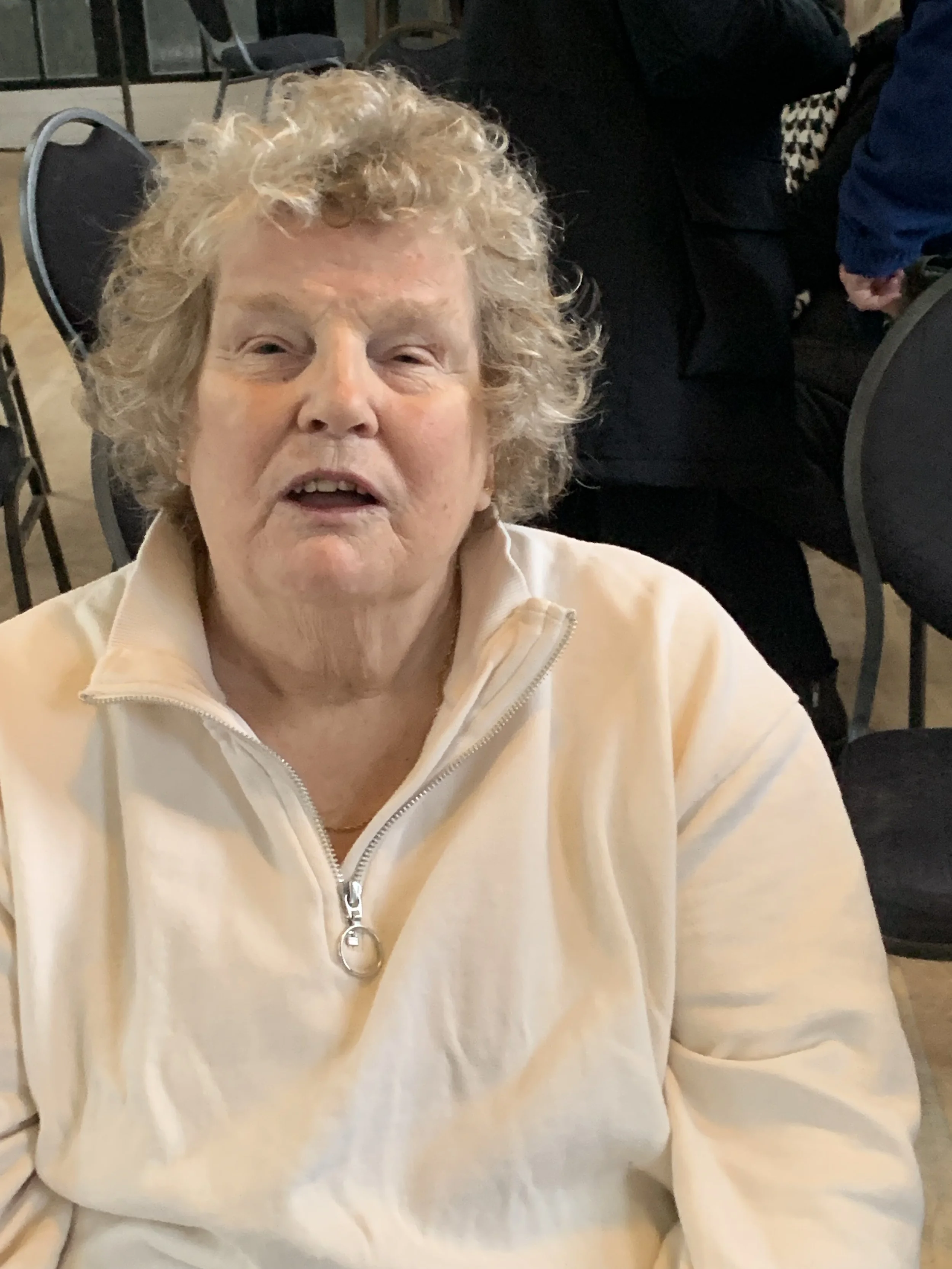 An elderly woman with curly, short, gray hair and light skin, wearing a light-colored zip-up top, sitting indoors among other people and chairs.