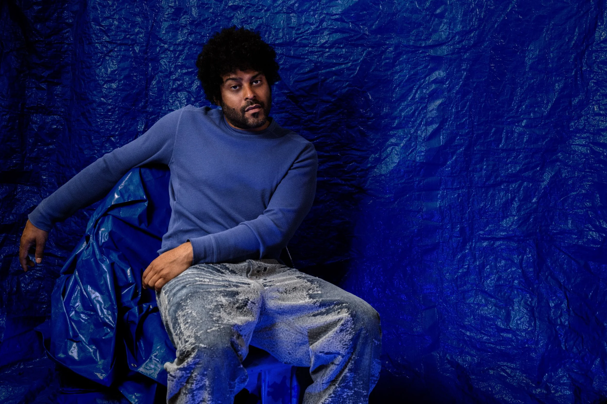 A man with curly black hair and a beard sits against a crinkled blue background. He is wearing a blue long-sleeve shirt and light-colored, paint-splattered pants.