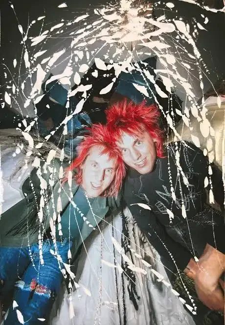 Two young people with bright red hair, smiling in a dark room with balloon-like decorations and white streamers overhead.