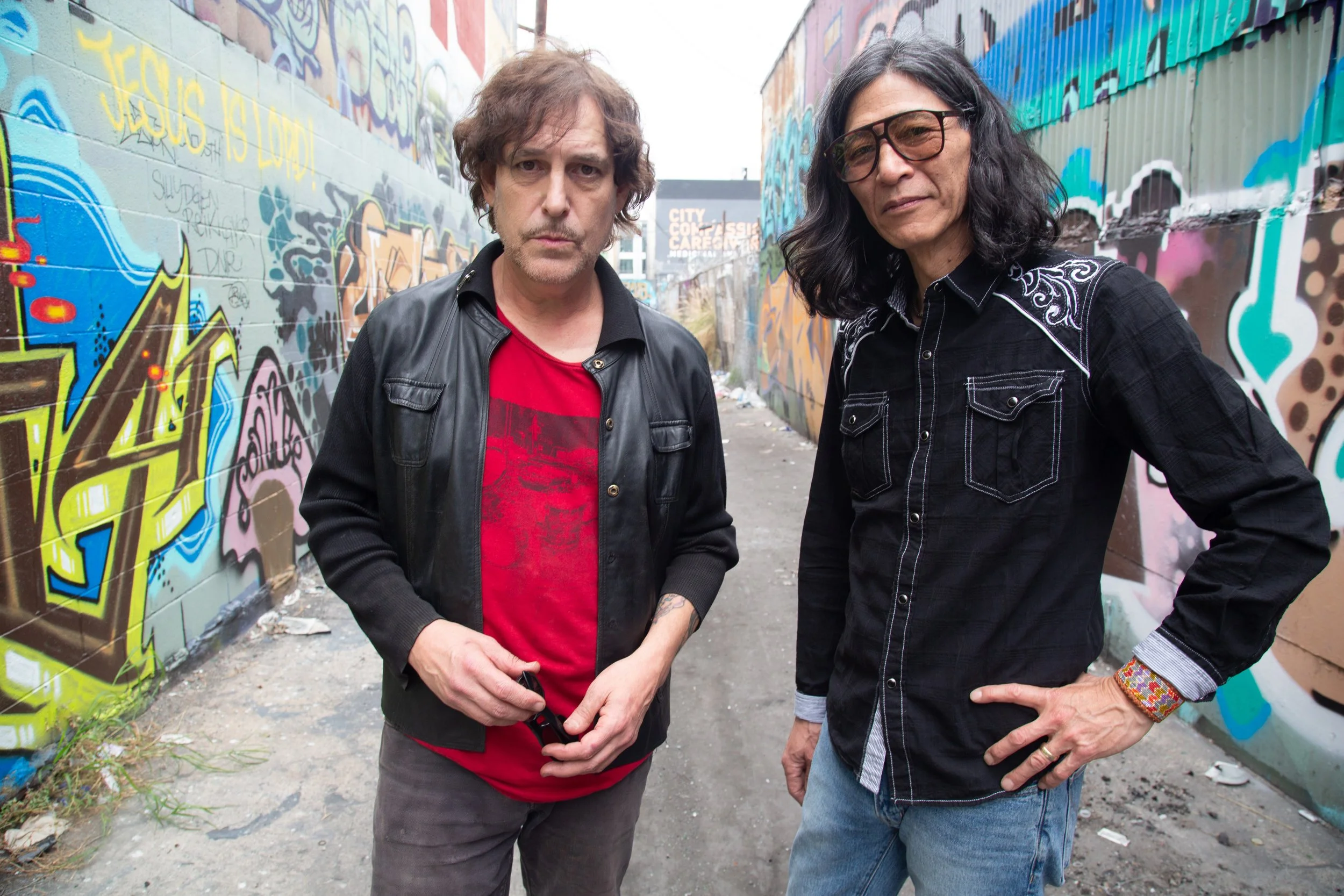 Two men stand in an alleyway with colorful graffiti on the walls. The man on the left has curly brown hair, wears a black leather jacket over a red T-shirt, and has a serious expression. The man on the right has long black hair, wears glasses, a black shirt with white embroidery, and has one hand on his hip.