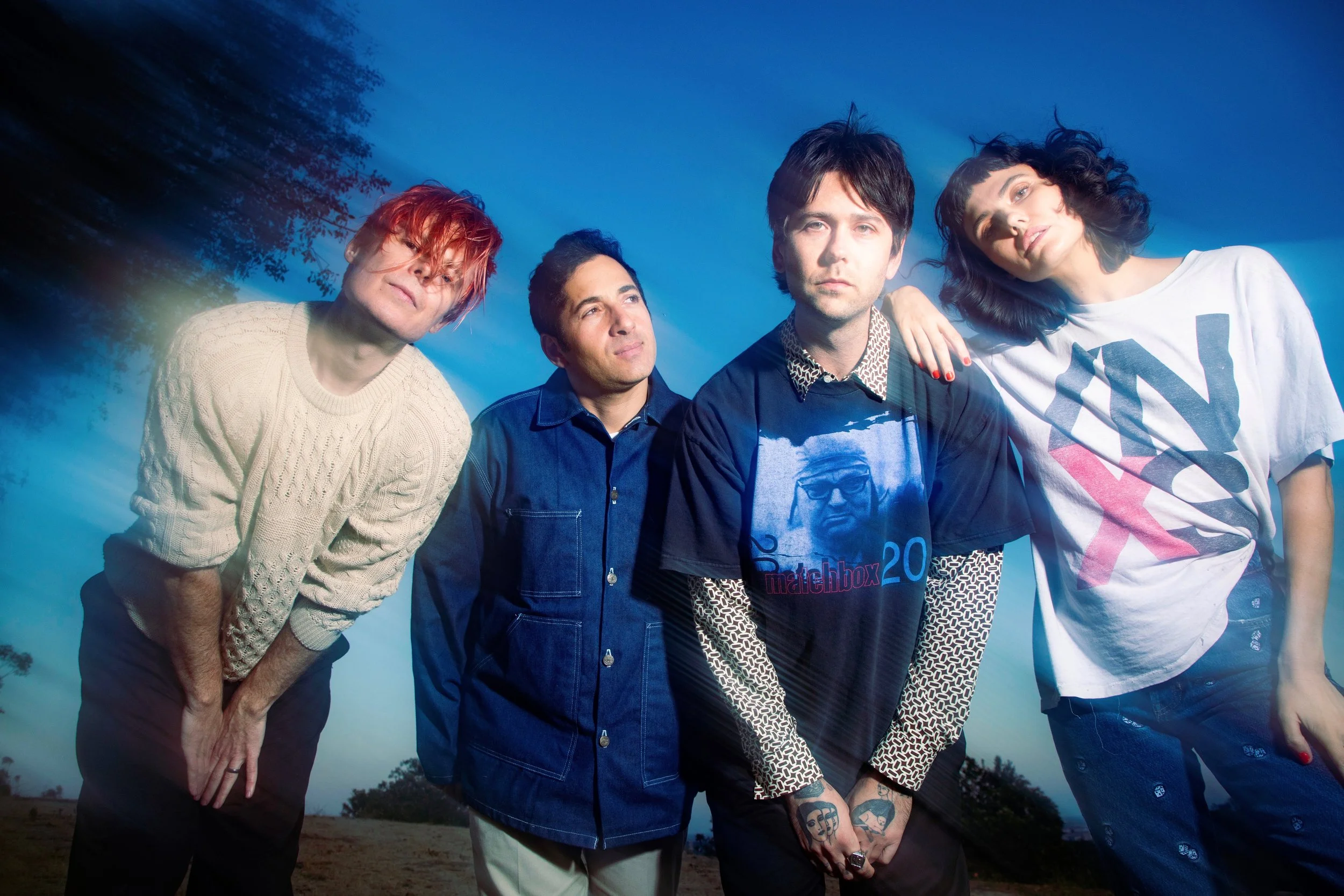 A group of four young adults posing outdoors during sunset or twilight, with trees and a clear sky in the background.