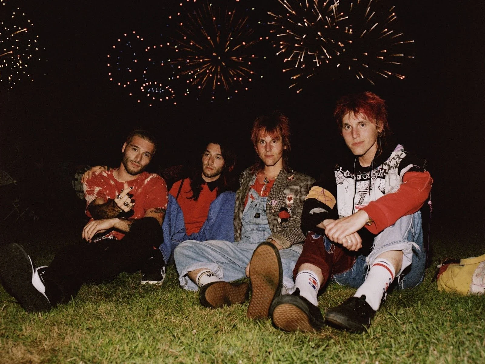 A group of four young men sitting on grass at night, with fireworks in the sky behind them, celebrating or enjoying an event.
