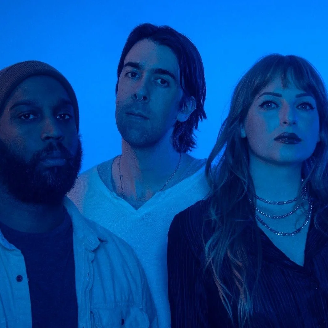 Three people standing against a blue background, looking at the camera.