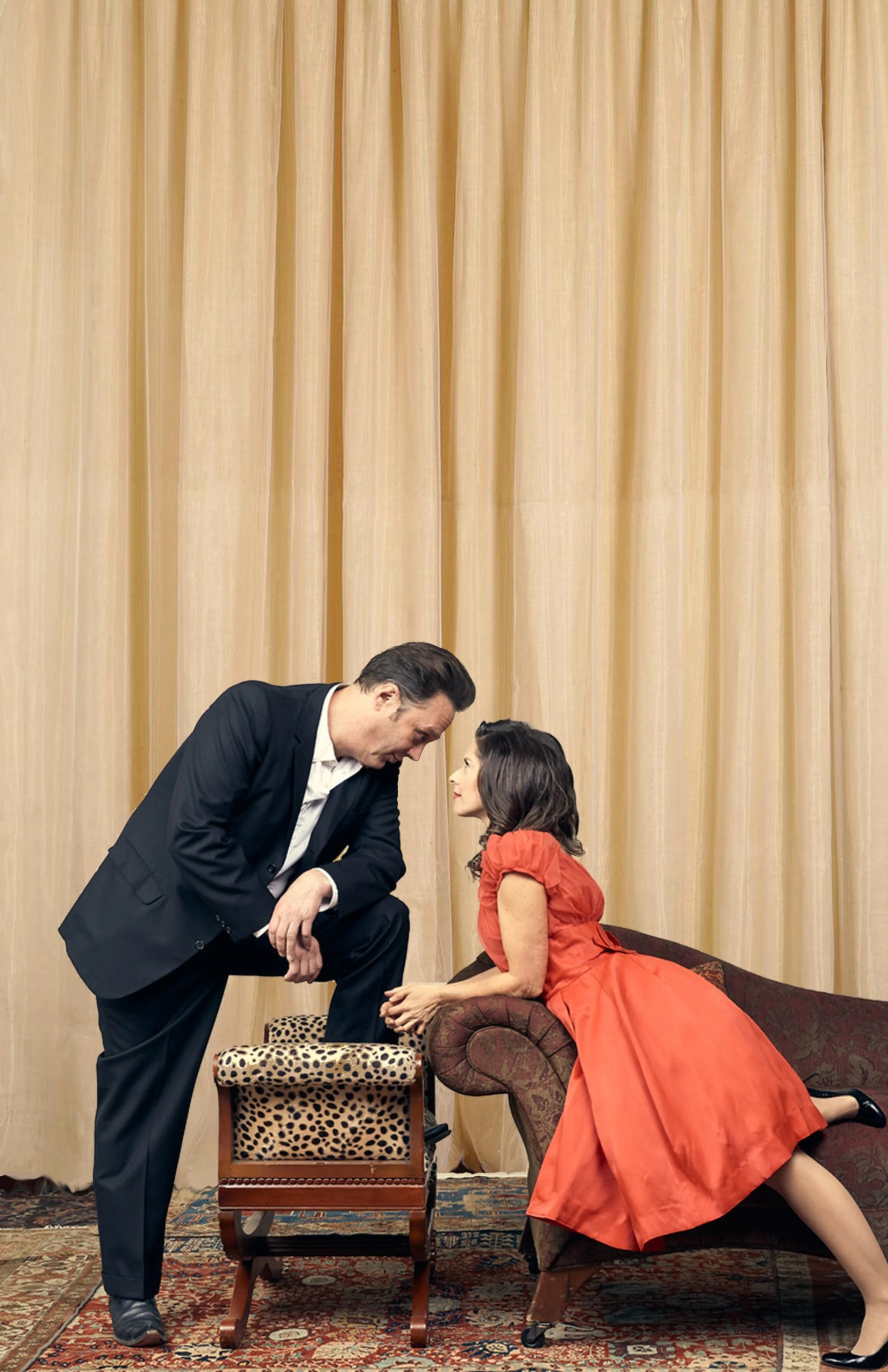 A man and a woman are engaged in an intense conversation in a vintage room with beige curtains and a patterned rug. The man is standing, leaning forward with one foot on a leopard-print rolling stool, while the woman is reclining on a dark, patterned chaise. She is dressed in a bright red dress and the man is dressed in a black suit.