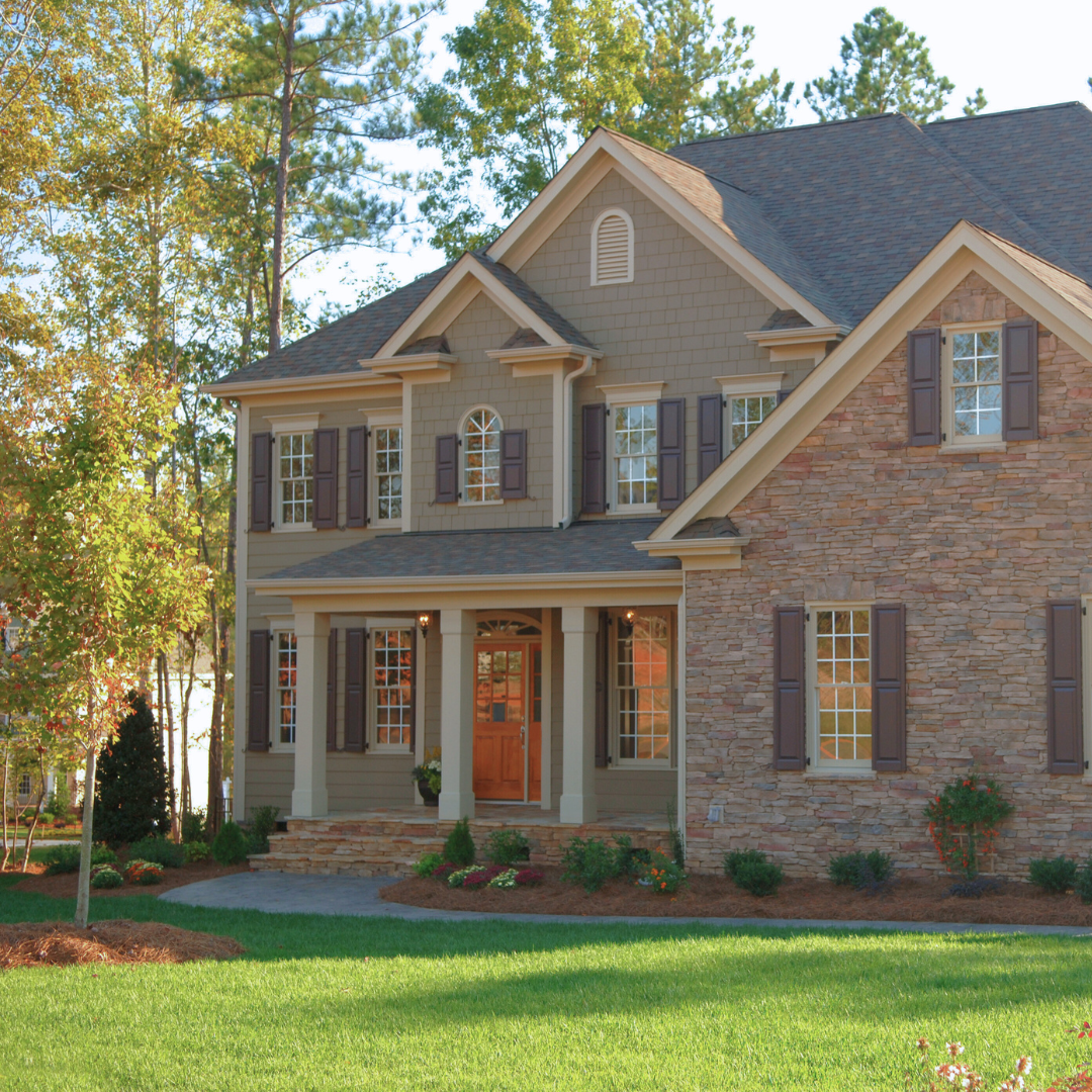 A two-story suburban house with beige siding, stone accents, and dark shutters, surrounded by a landscaped yard with green grass, trees, and bushes.