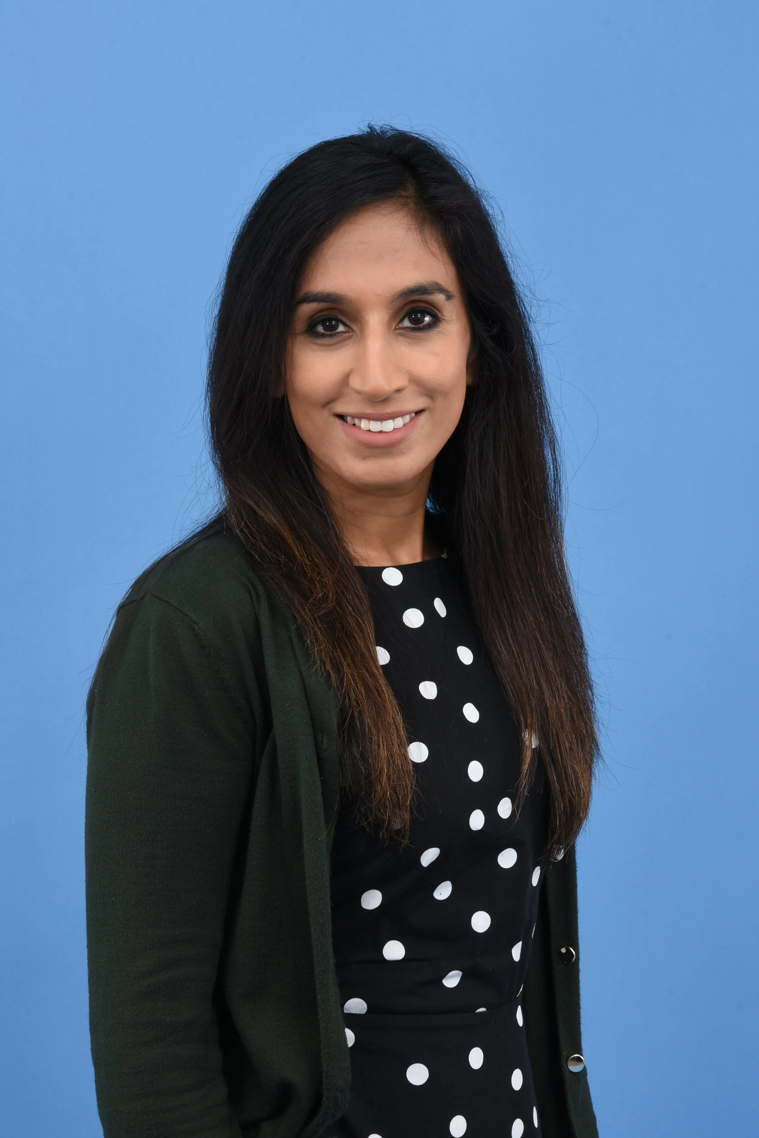 A woman with long dark hair, wearing a black dress with white polka dots and a dark green cardigan, smiling, against a blue background.