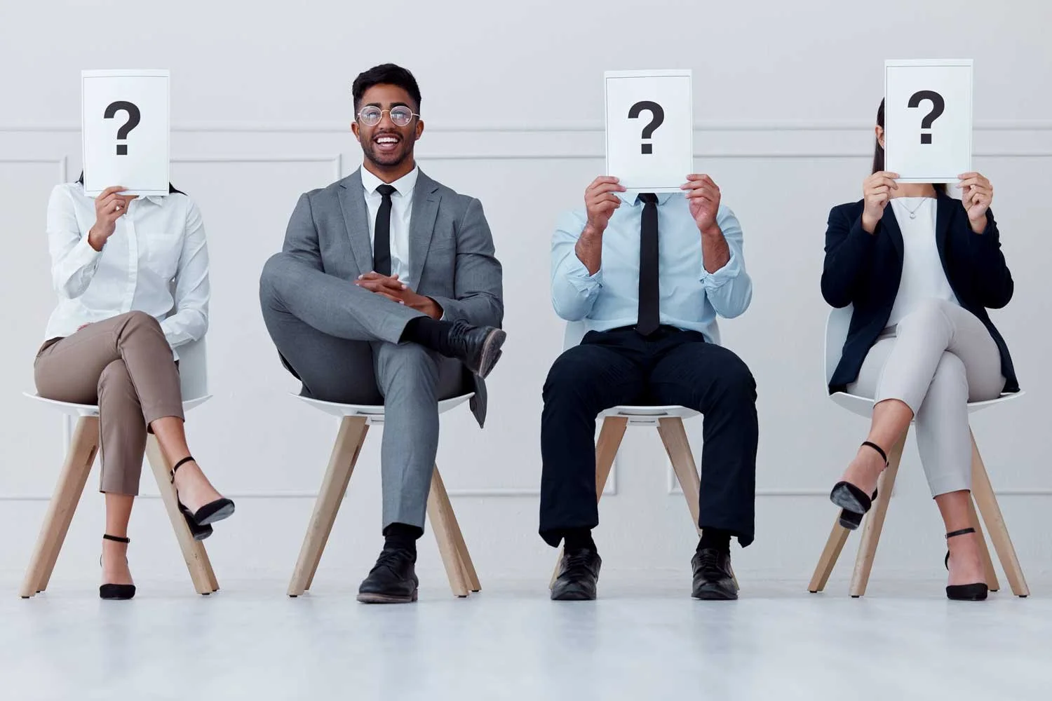 Three seated people cover faces with cards that have question marks and one seated man smiling