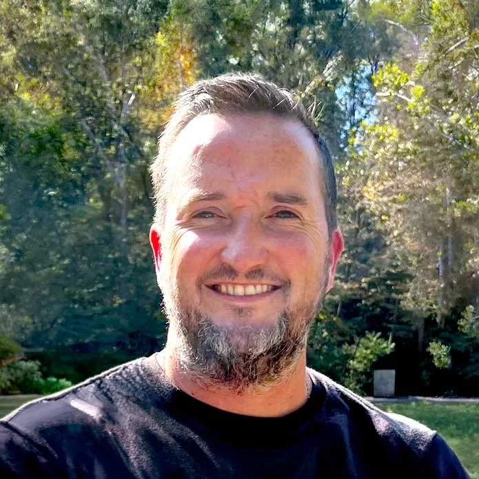 A man smiling outdoors with trees and sunlight in the background.