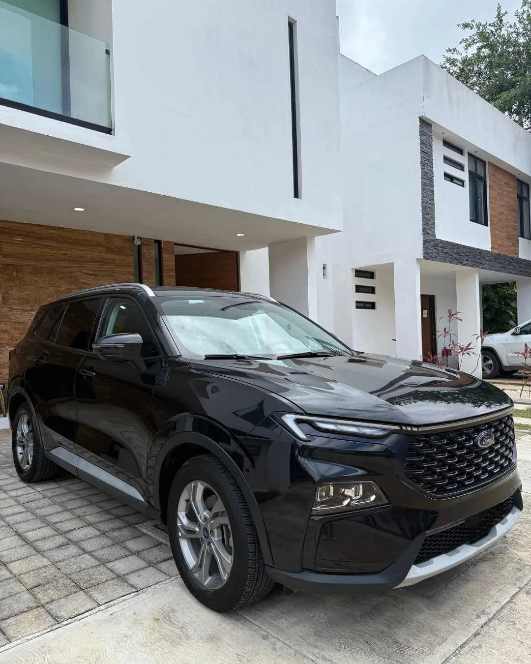 Coche negro en la entrada de una casa moderna de dos pisos, con detalles en madera y paredes blancas.