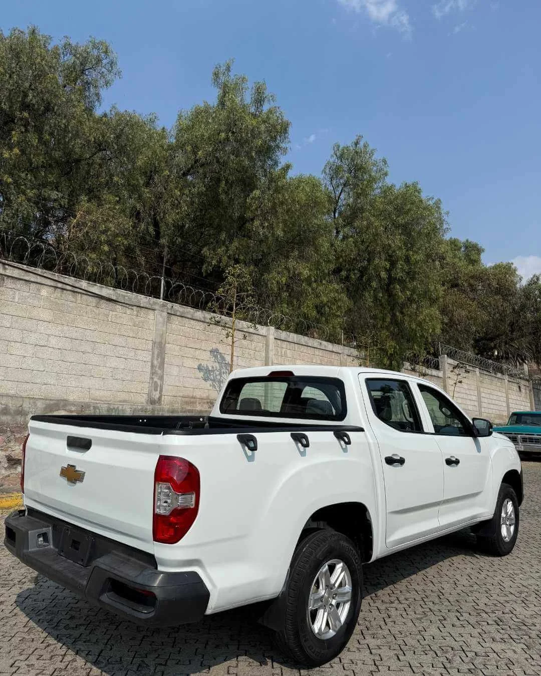 Camioneta blanca de doble cabina con logo de Chevrolet estacionada en una calle con muro de bloque y arbustos verdes al fondo.