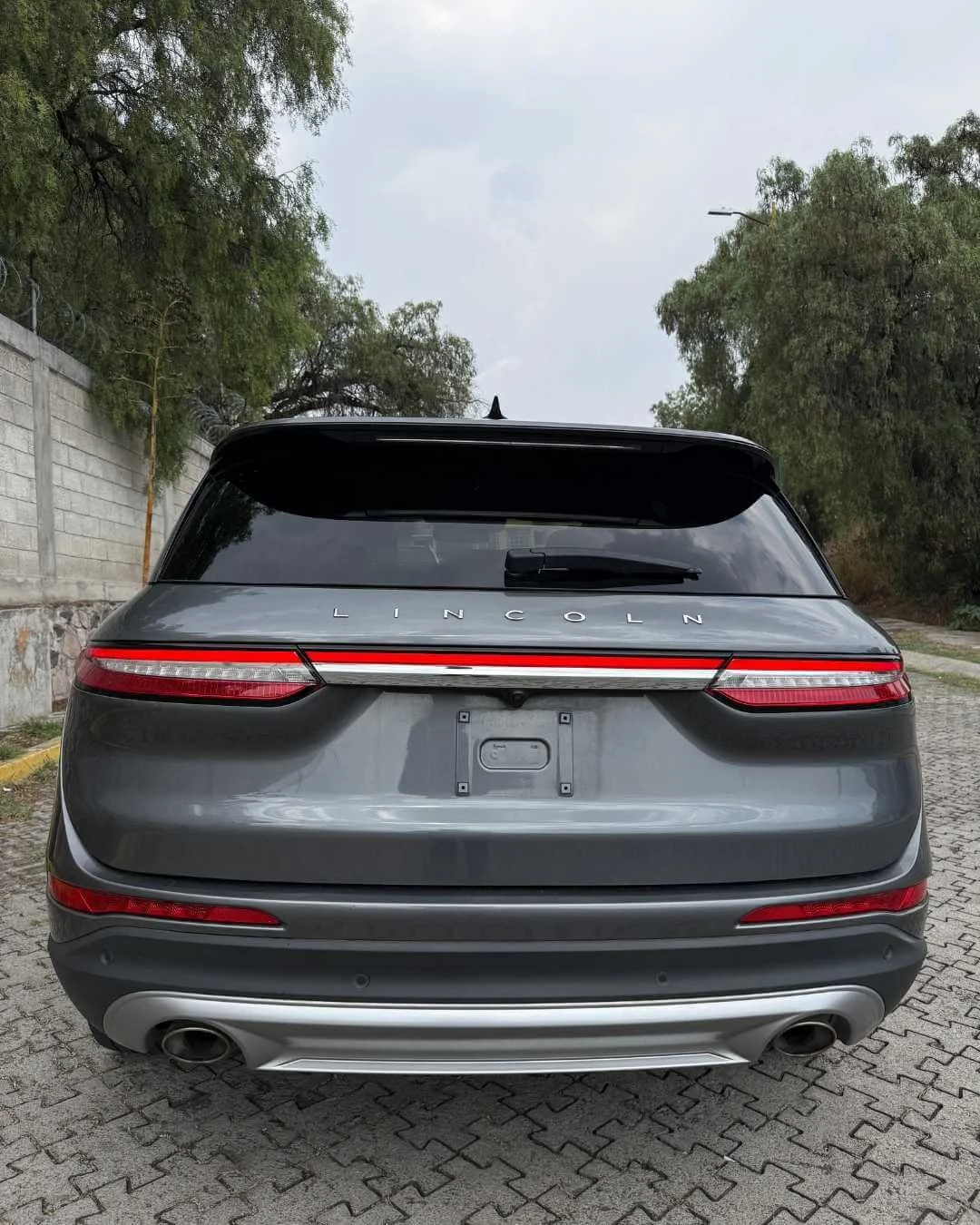 Vista trasera de un auto gris marca Lincoln, sin placa, estacionado en una calle con árboles y un muro de fondo.