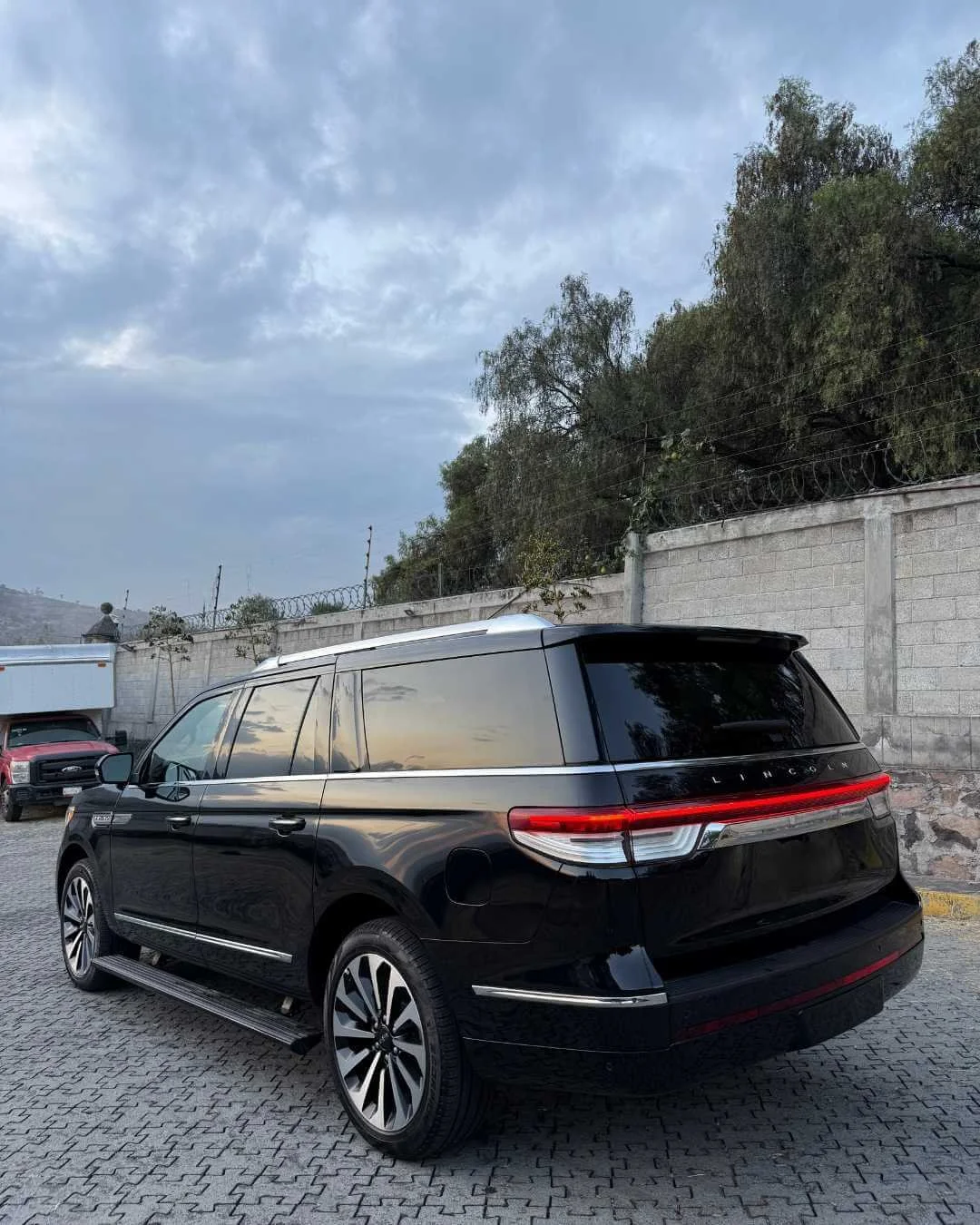 Auto negro tipo SUV estacionado en un área pavimentada con paredes de ladrillo y árboles al fondo, bajo un cielo nublado.
