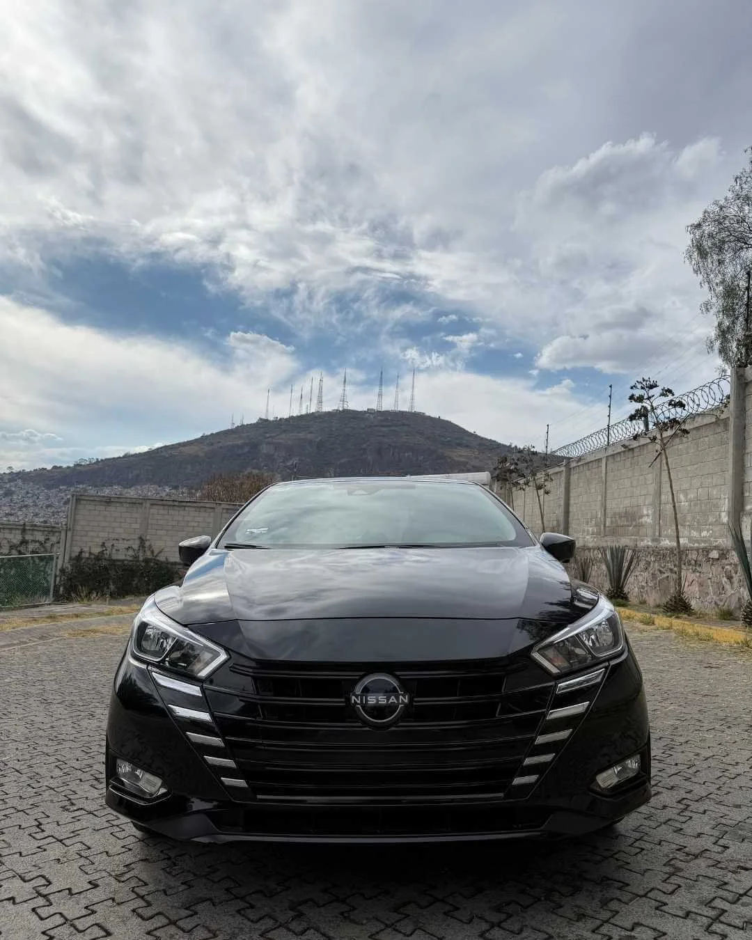 Auto negro Nissan estacionado en un área pavimentada con muro de ladrillo y montaña con antenas en el fondo, cielo nublado.