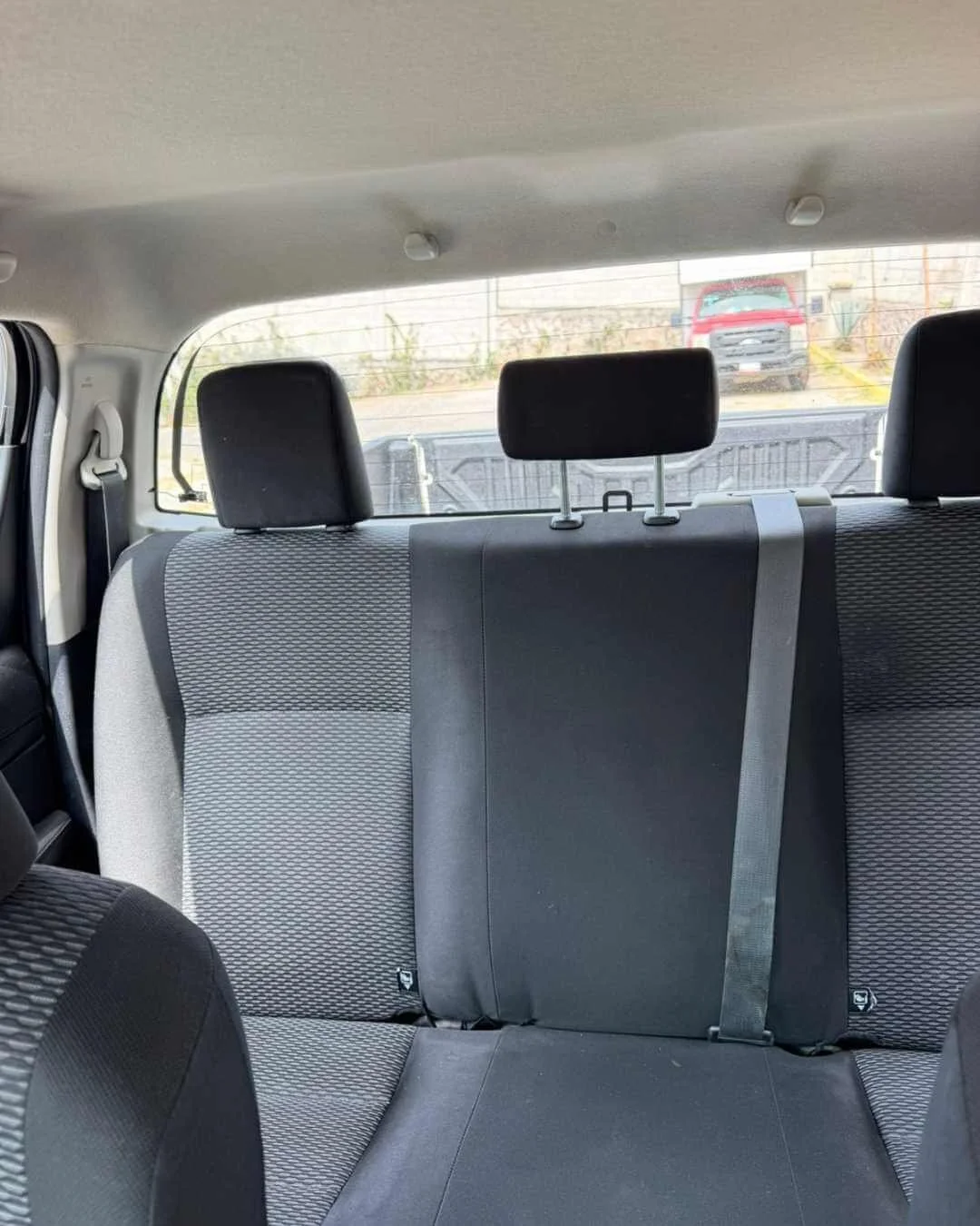 Asiento trasero de un vehículo con el respaldo, cinturón de seguridad y cabeceras visibles, y una ventana trasera con una camioneta roja y una cerca en el fondo.