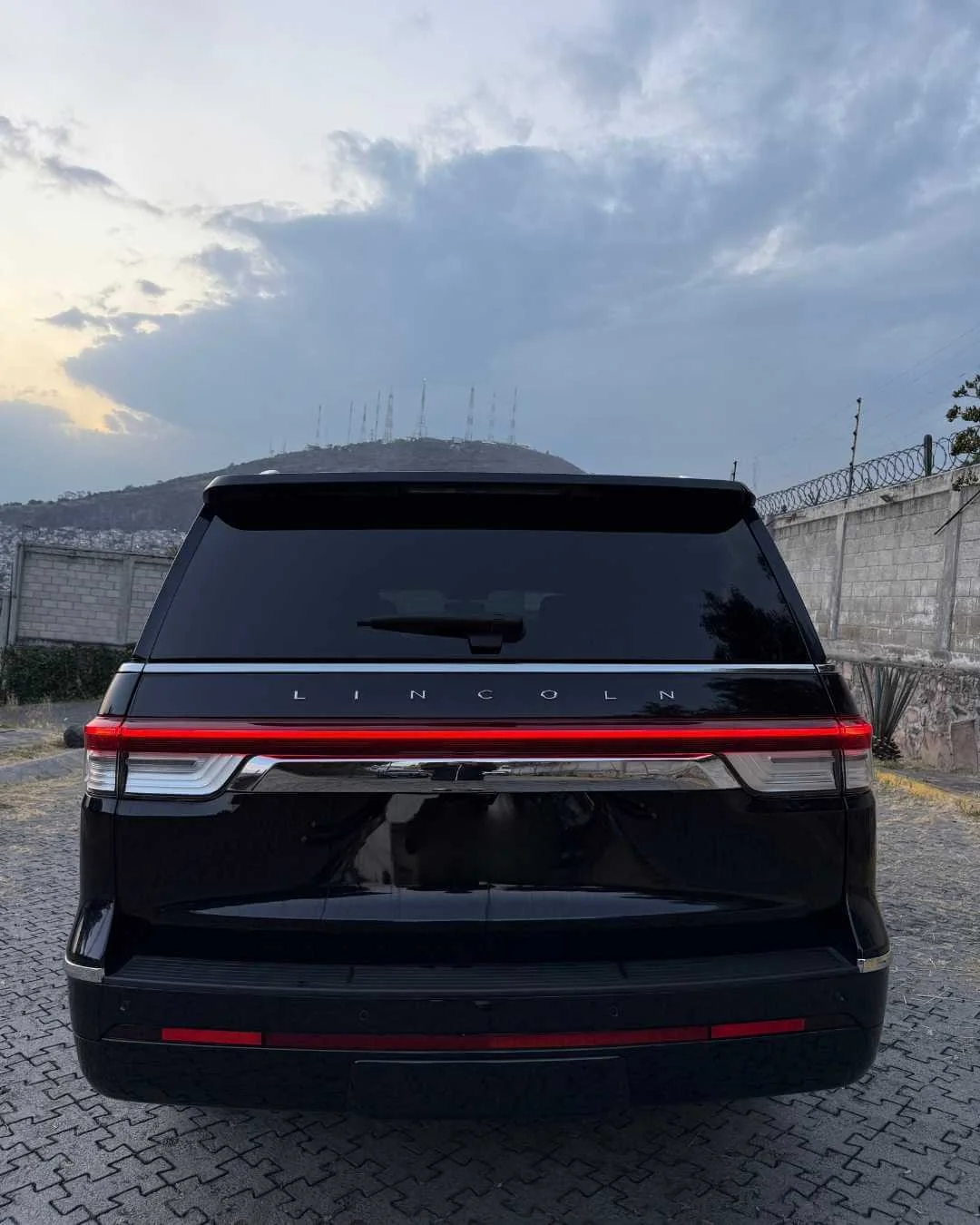 Respaldo de un automóvil Lincoln negro con luz trasera en un estacionamiento con muro de piedra y montaña en el fondo bajo cielo nublado.