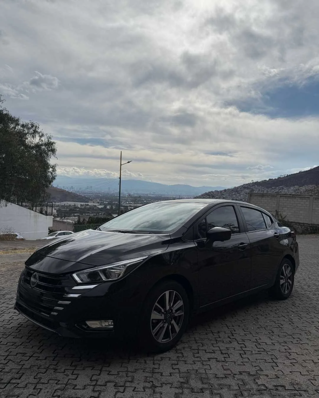 Auto negro estacionado sobre pavimento de adoquines con montañas y cielo nublado al fondo.