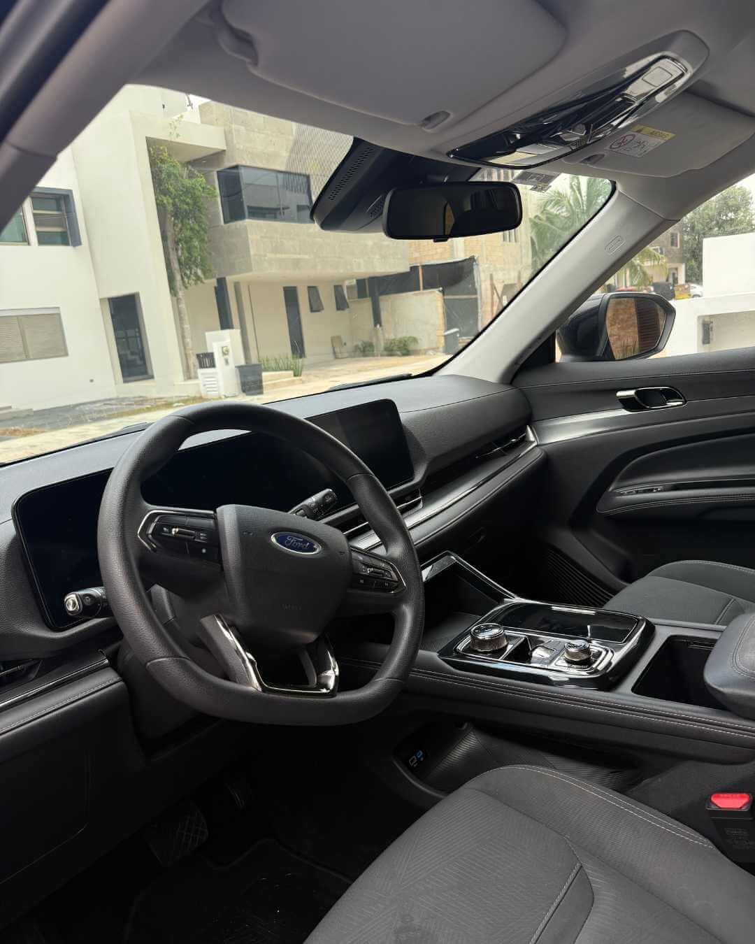 Interior de un vehículo Ford con volante, tablero digital, pantalla central, control de transmisión y asientos grises, con vista a una construcción moderna y una calle en el fondo.