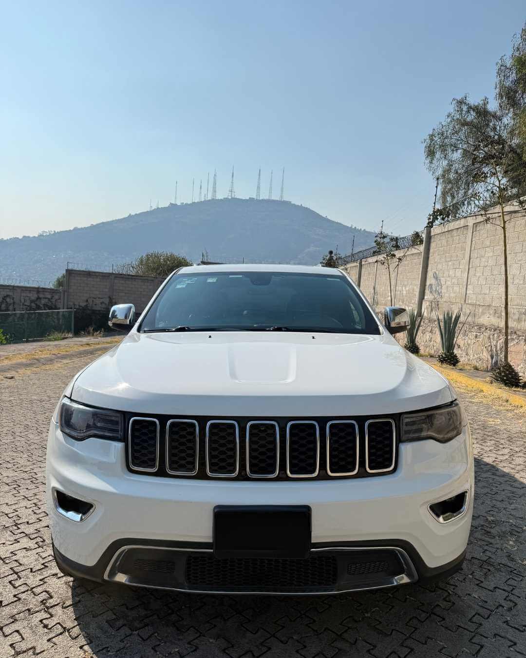 Auto blanco estacionado en una calle adoquinada con colinas y antenas en el fondo.