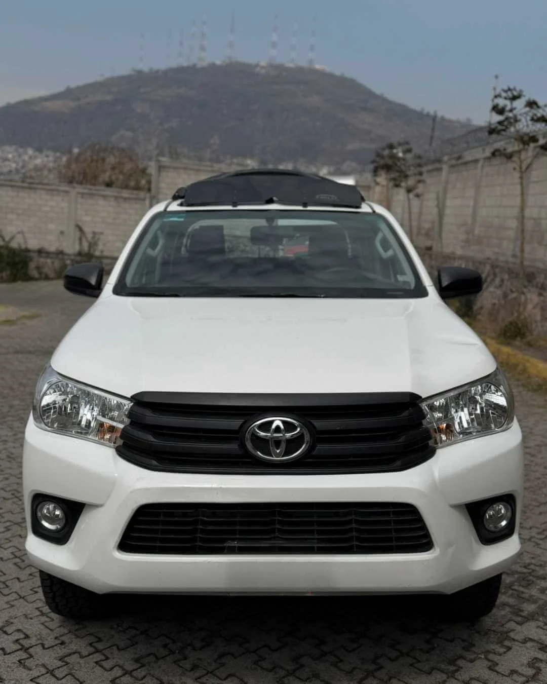 Auto SUV, color blanco, en un estacionamiento, con colina y antenas en el fondo.