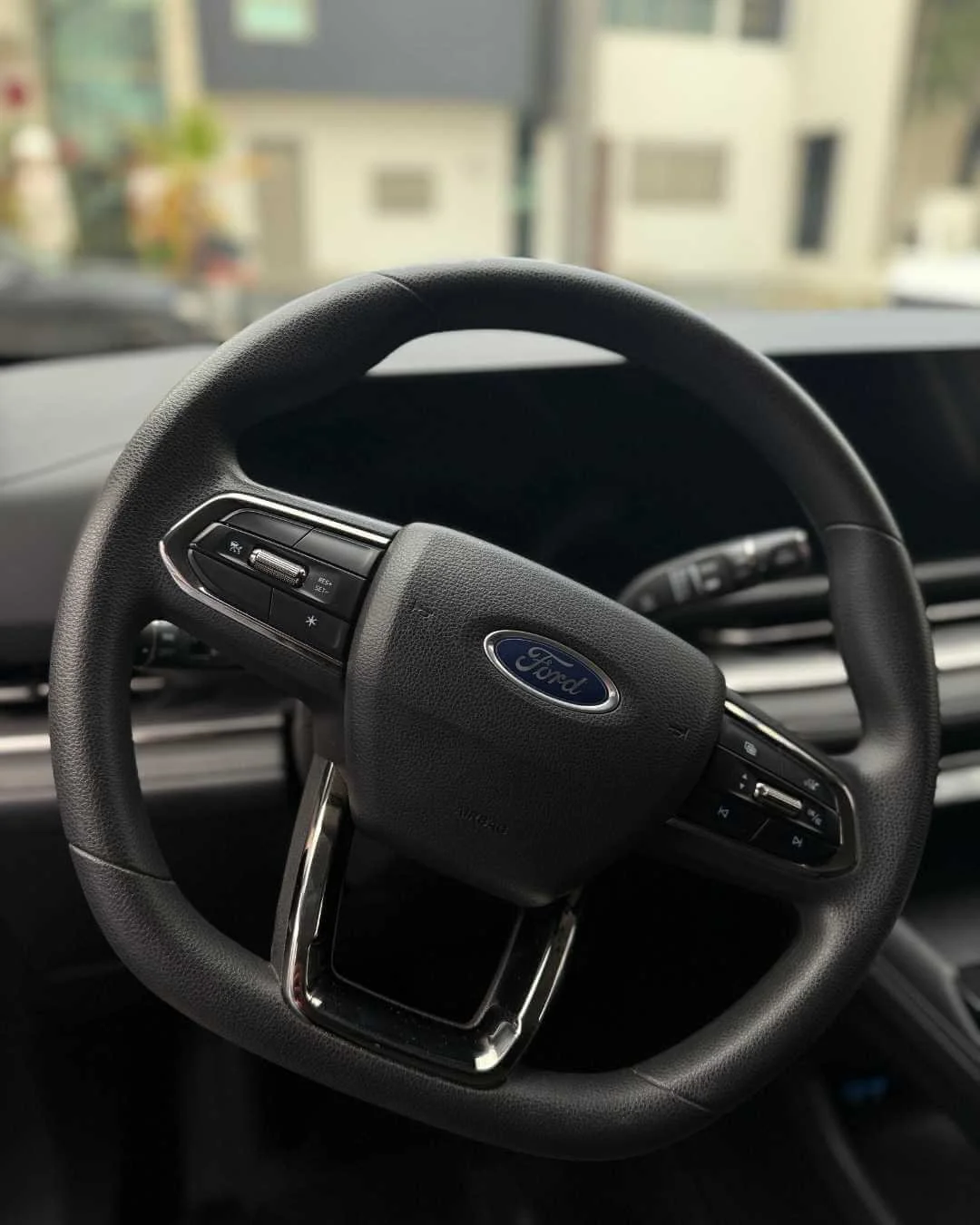 Volante de coche marca Ford con controles en el lado izquierdo y derecho, fondo difuso del interior y exterior de edificios.